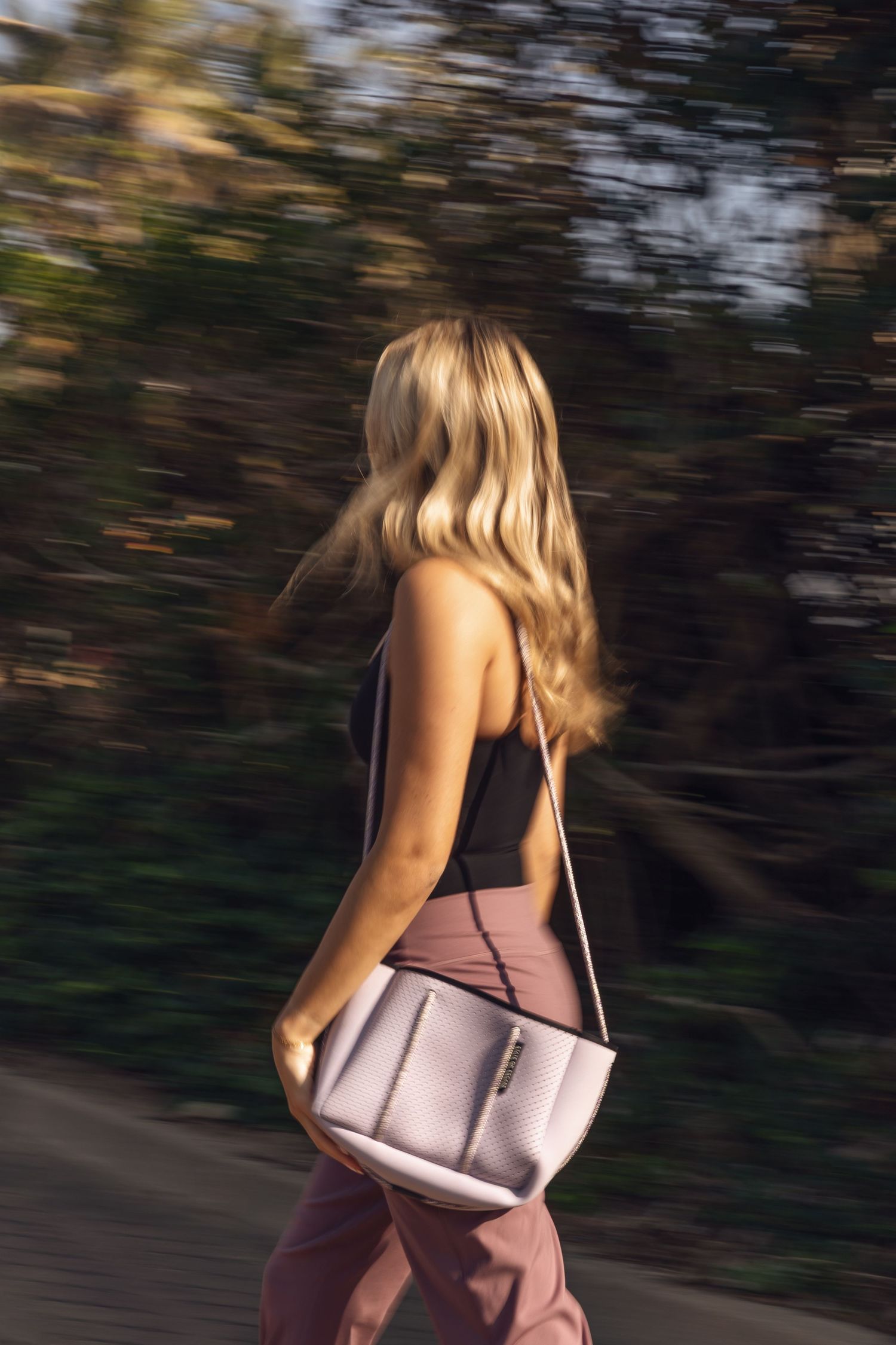 Person in motion wearing black tank top and brown pants walking with a beige shoulder bag against blurred background.