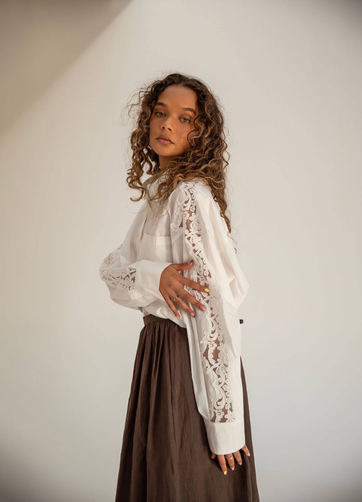 Woman in white lace blouse and brown maxi skirt poses sideways with curly hair against neutral background.