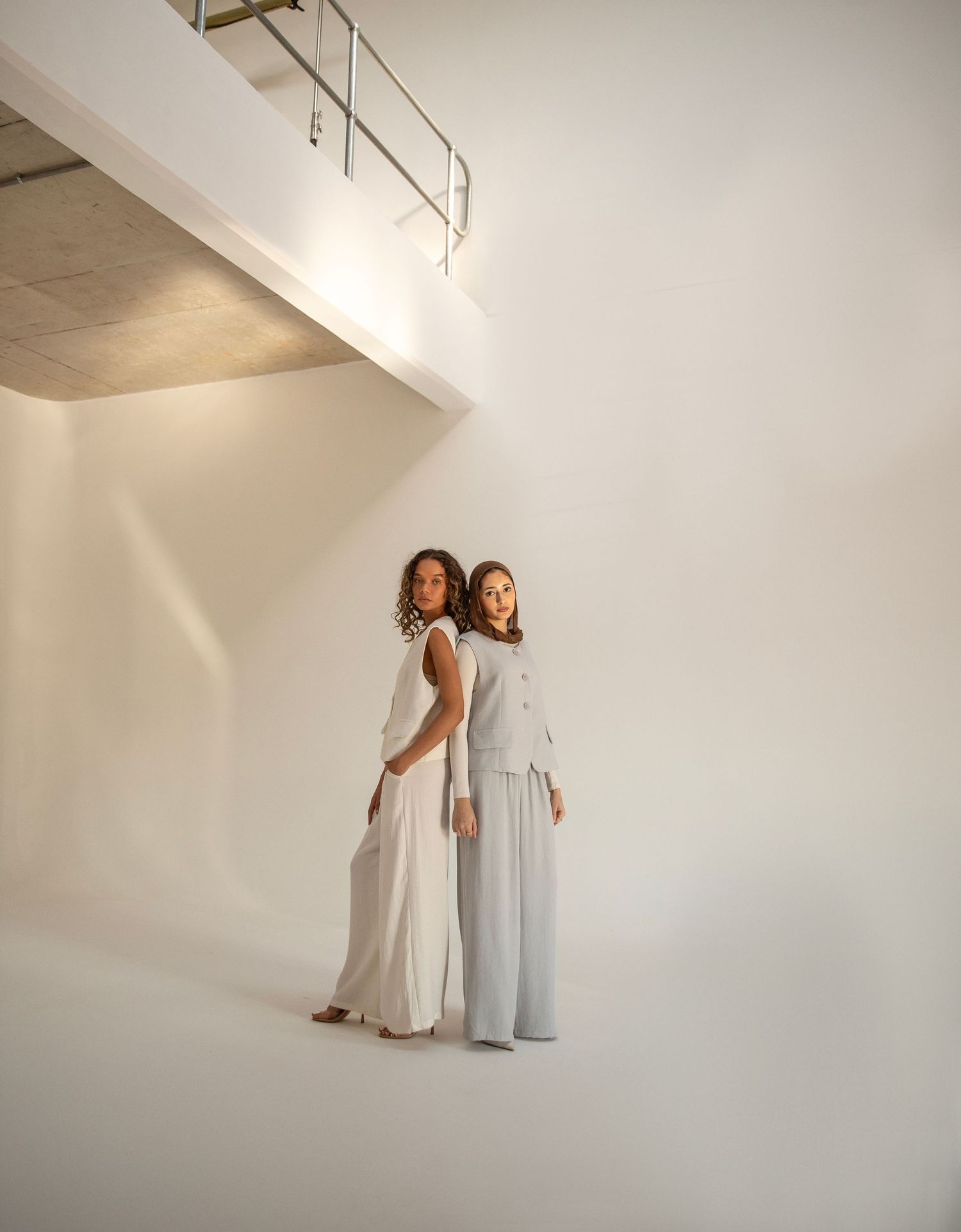 Two women in white and light gray outfits stand back-to-back in a minimalist white studio space.