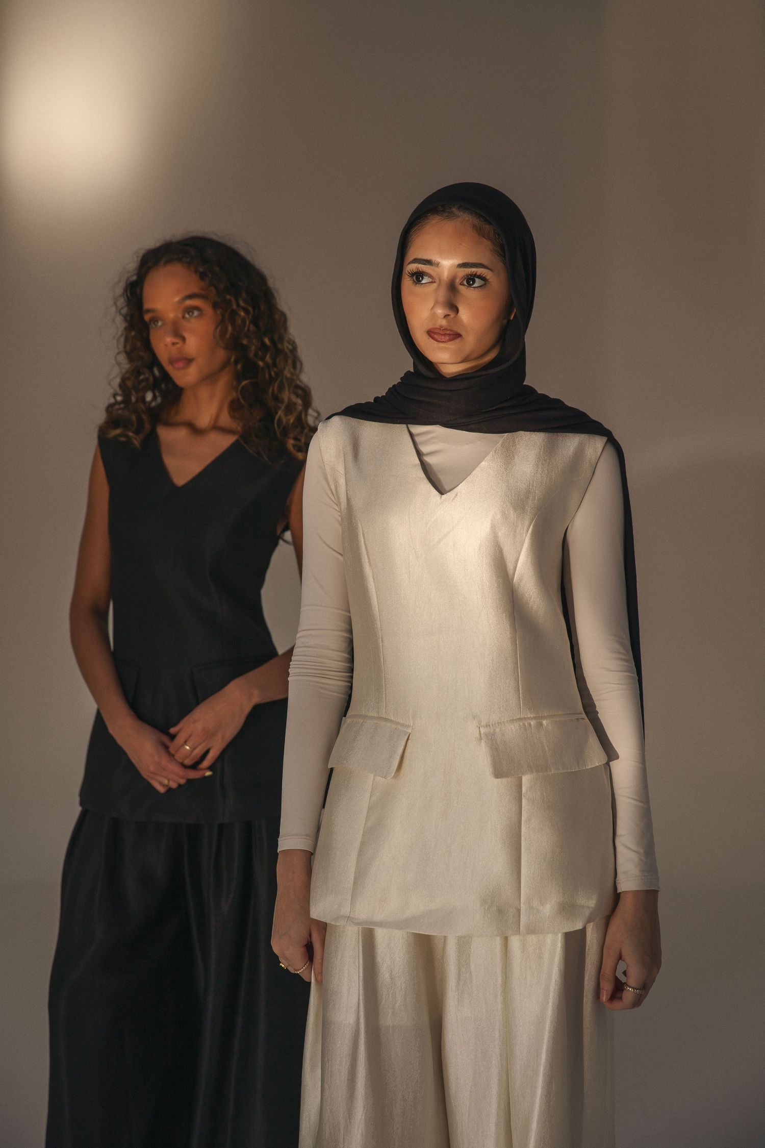 Two models in minimalist outfits, one in black sleeveless dress, one in white structured top with dark hijab.