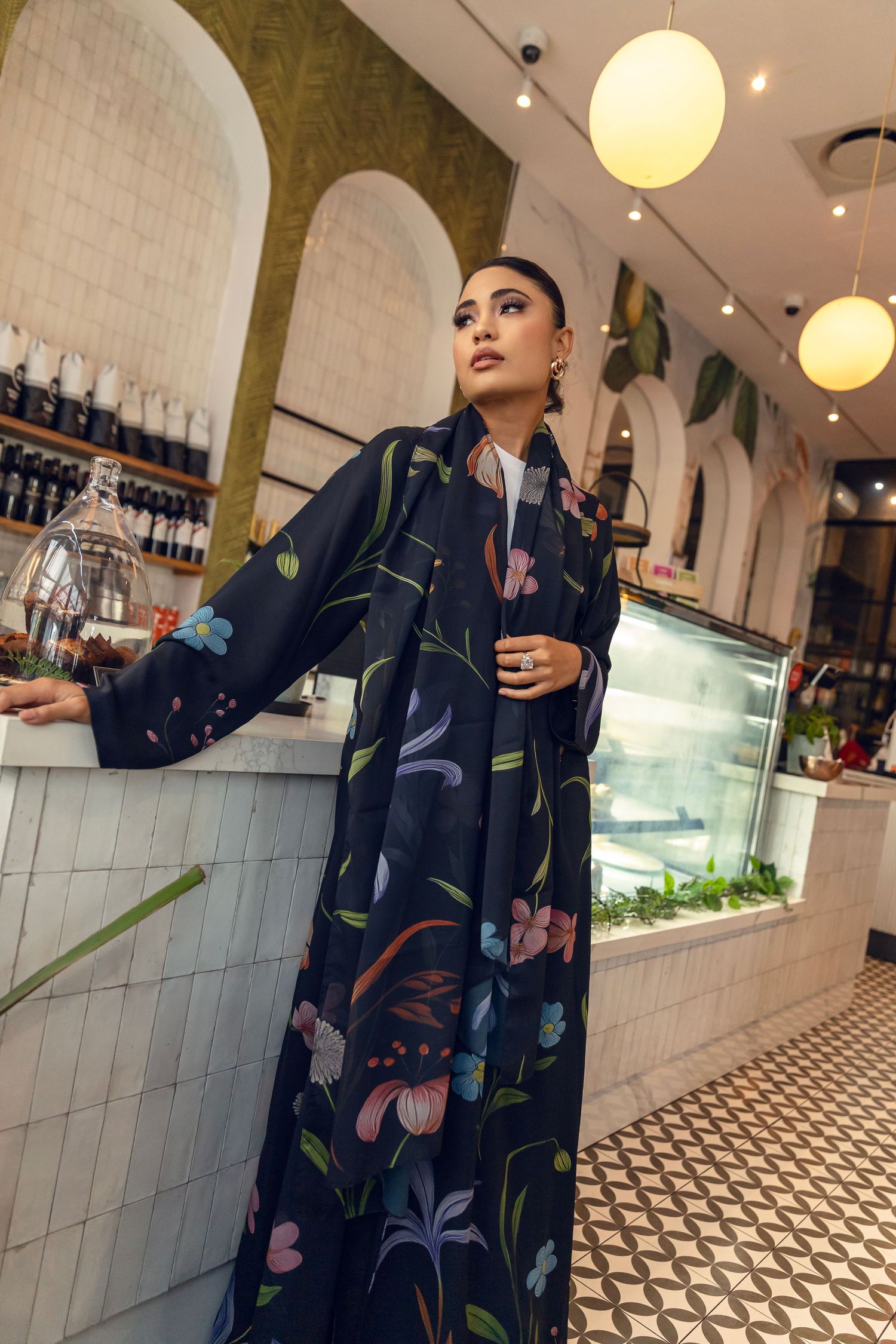 Fashion model poses in a floral print navy dress against modern restaurant interior with white tile walls and pendant lights.