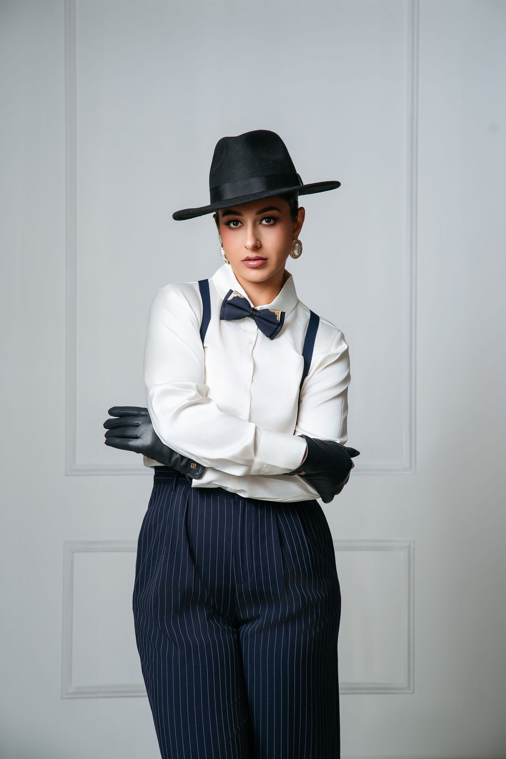 Woman in white shirt, black pinstripe pants, suspenders, bow tie, fedora hat, and leather gloves posing confidently.