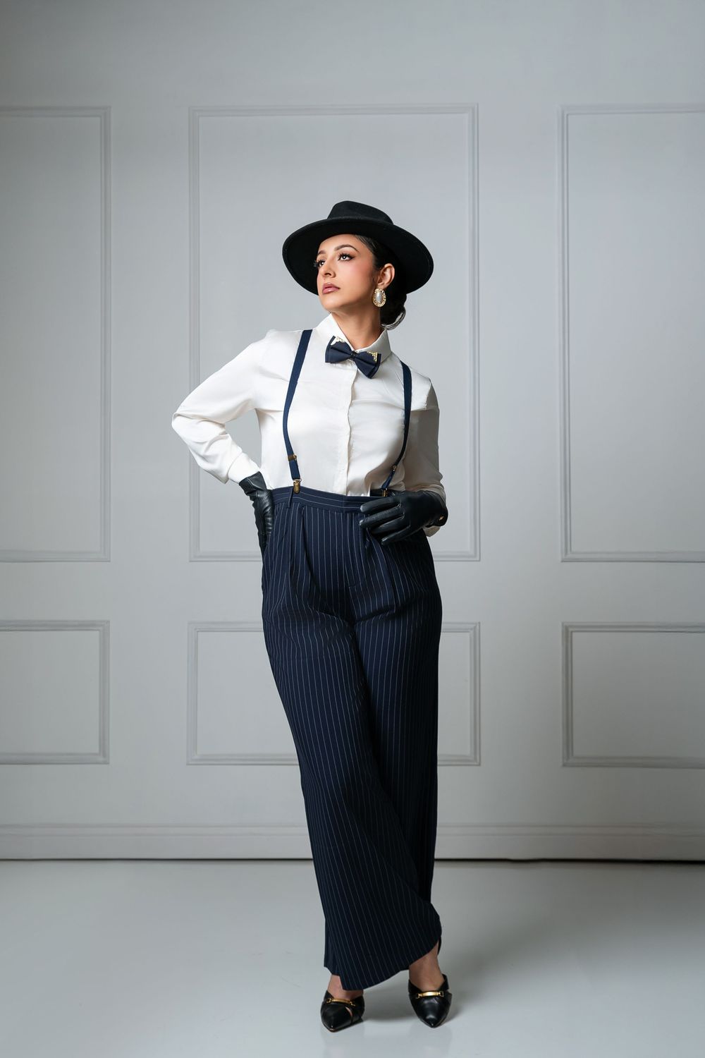 Woman in white shirt, suspenders, pinstripe trousers, black hat and gloves poses confidently in studio.