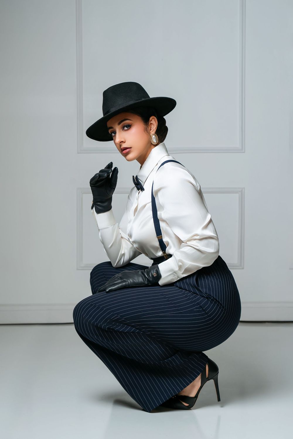 Elegant woman in pinstripe trousers, white blouse, suspenders, black gloves, and wide-brim hat posing in studio.