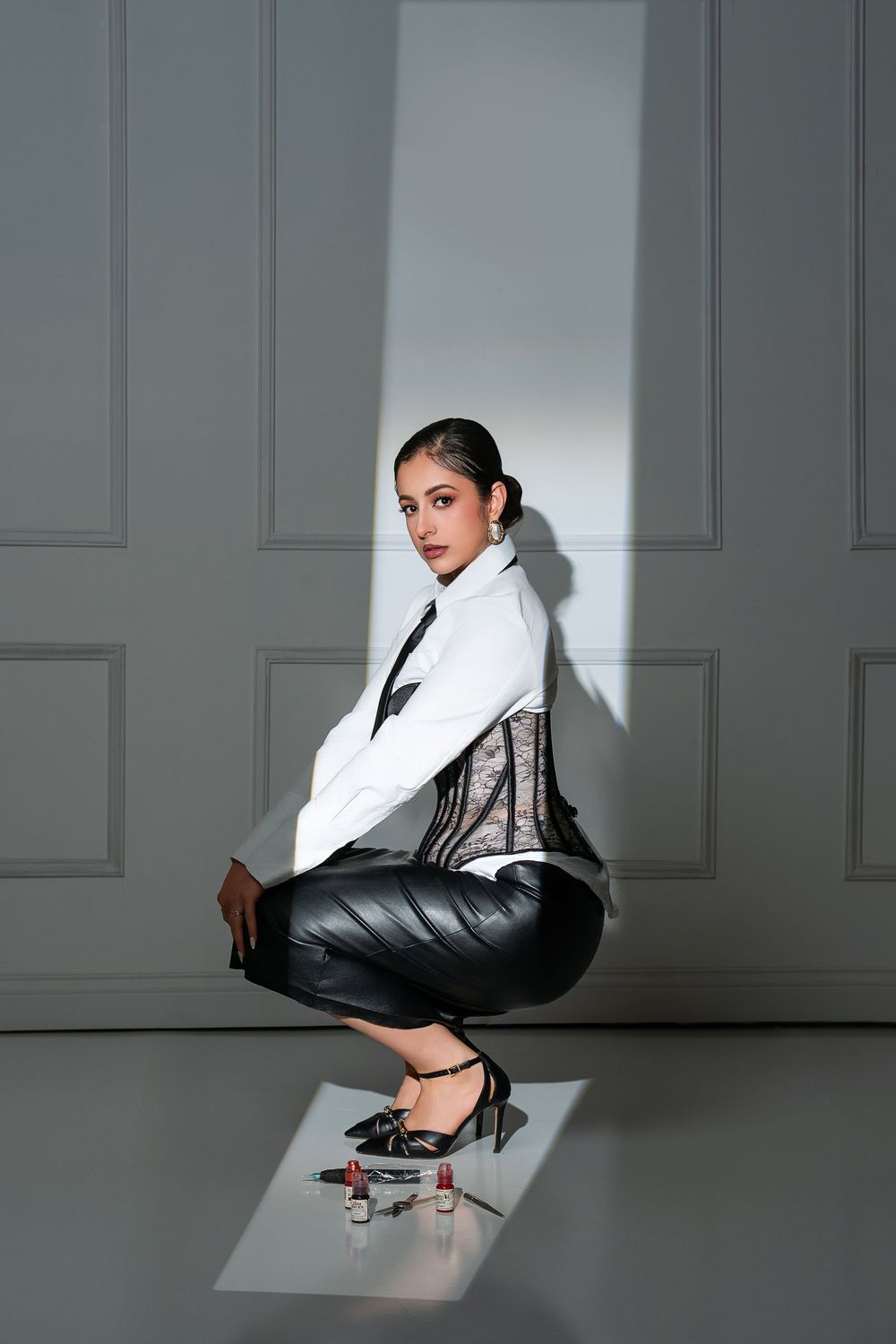 Woman in white blazer, lace corset, leather pants, and heels crouching elegantly against a white paneled wall.