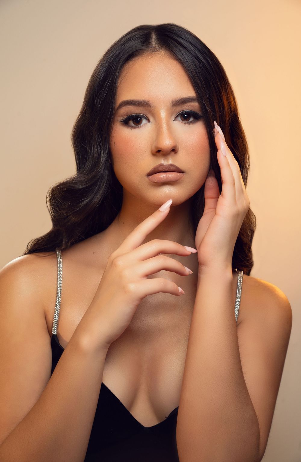 Young woman with wavy dark hair poses gracefully, wearing a black dress with rhinestone straps against a warm beige background.
