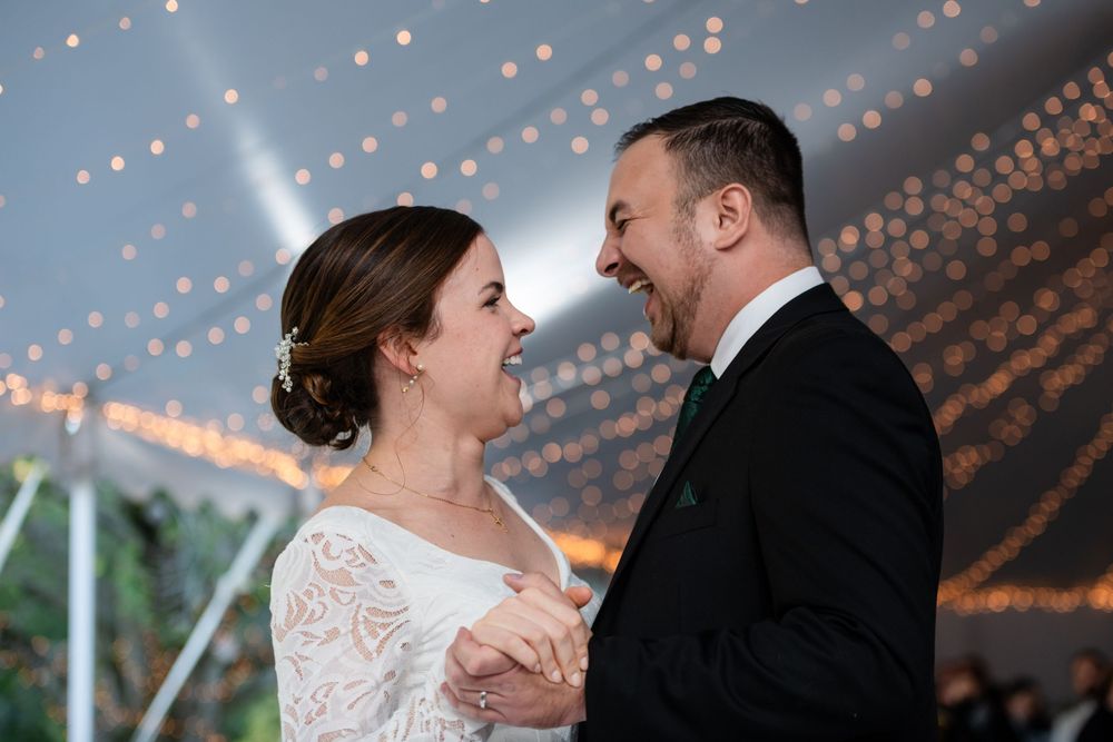 Newlyweds laughing together during their first dance beneath string lights at an outdoor wedding reception