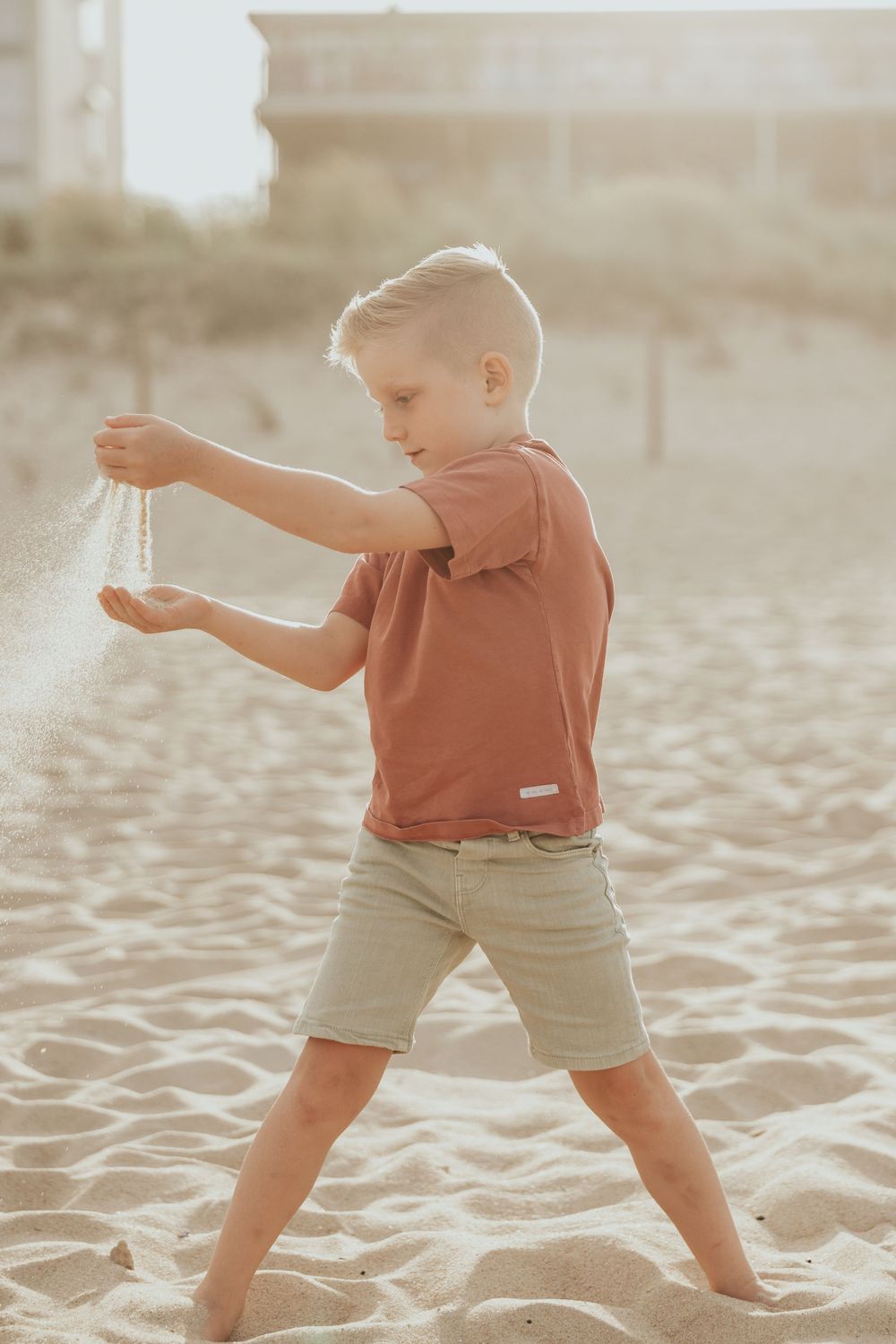 Child in coral colored shirt and khaki shorts playing and throwing sand at the beach during golden hour.