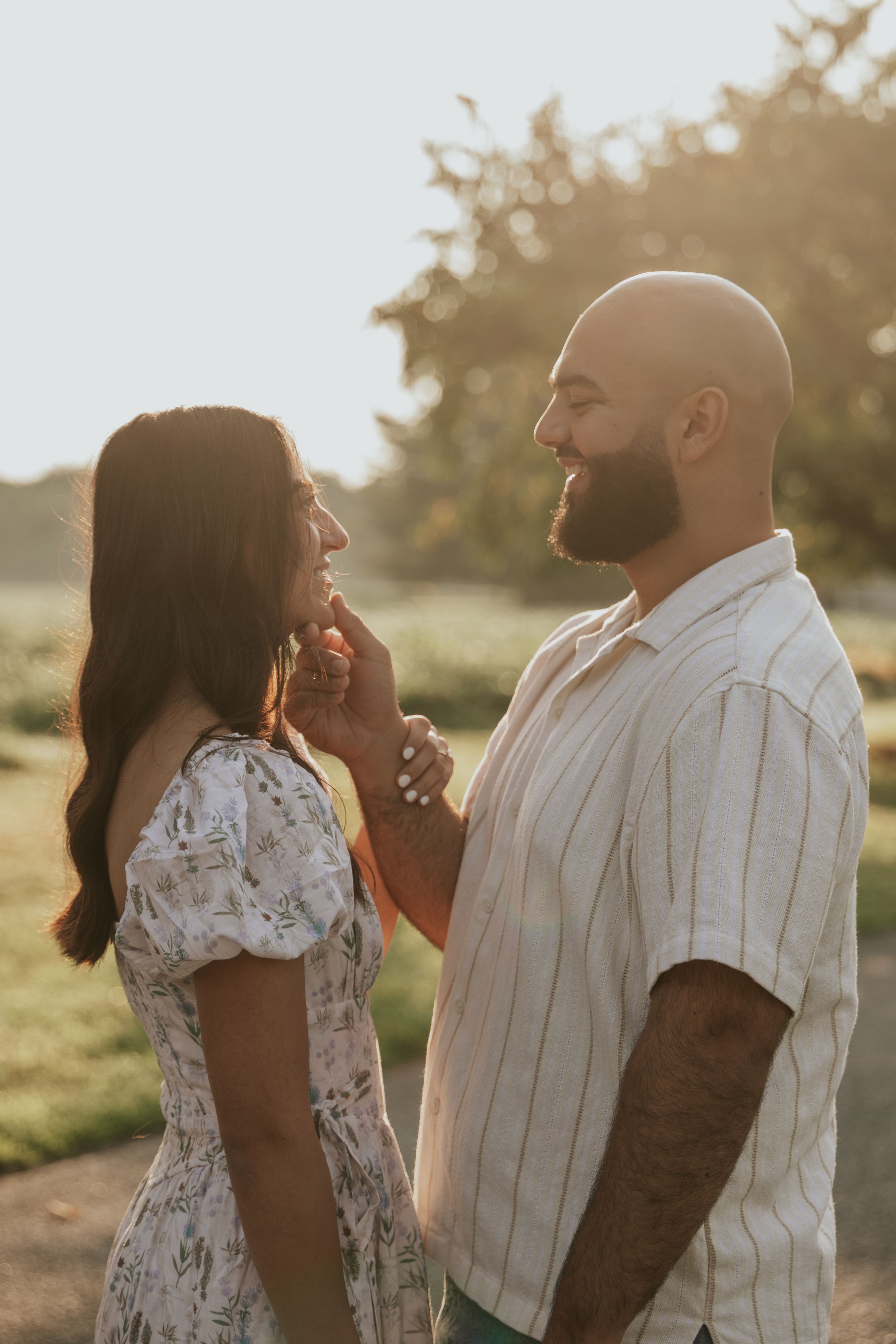 A couple shares an intimate moment in a sun-lit field during golden hour while wearing white and floral clothing.