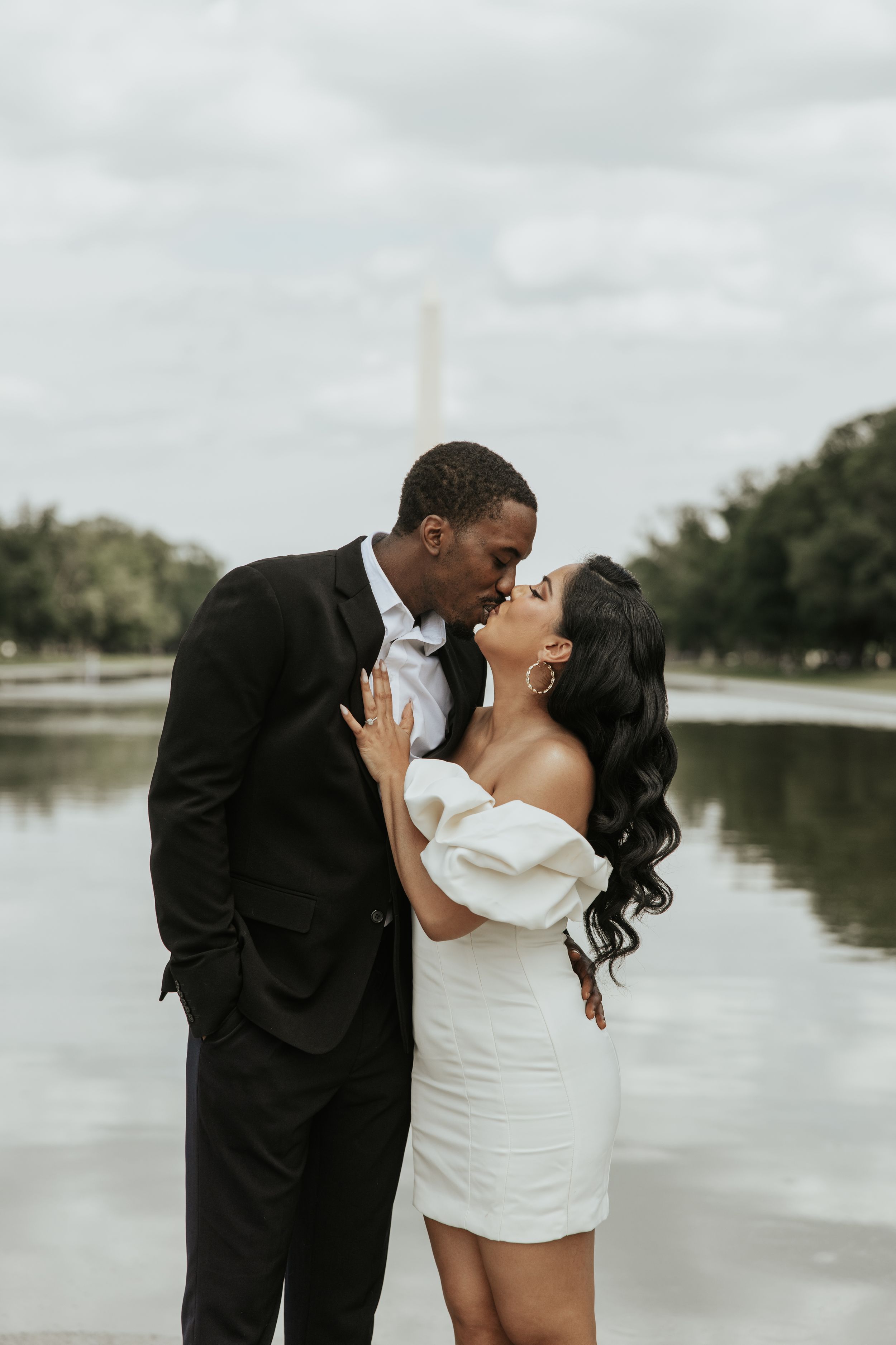 A romantic kiss between a couple in formal attire by a reflecting pool.