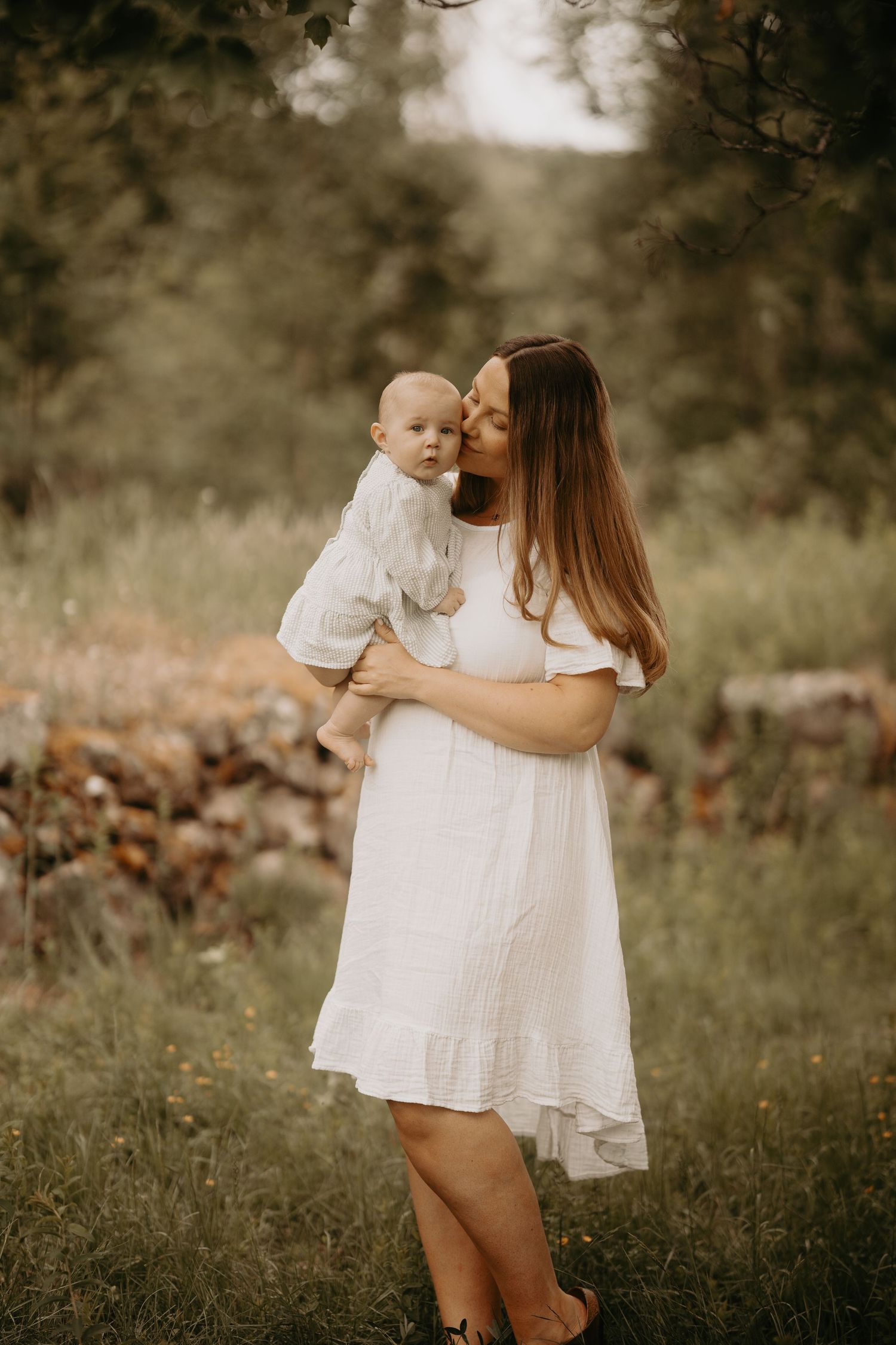 A tender outdoor portrait of mother and baby in white dresses standing in a sunlit meadow.