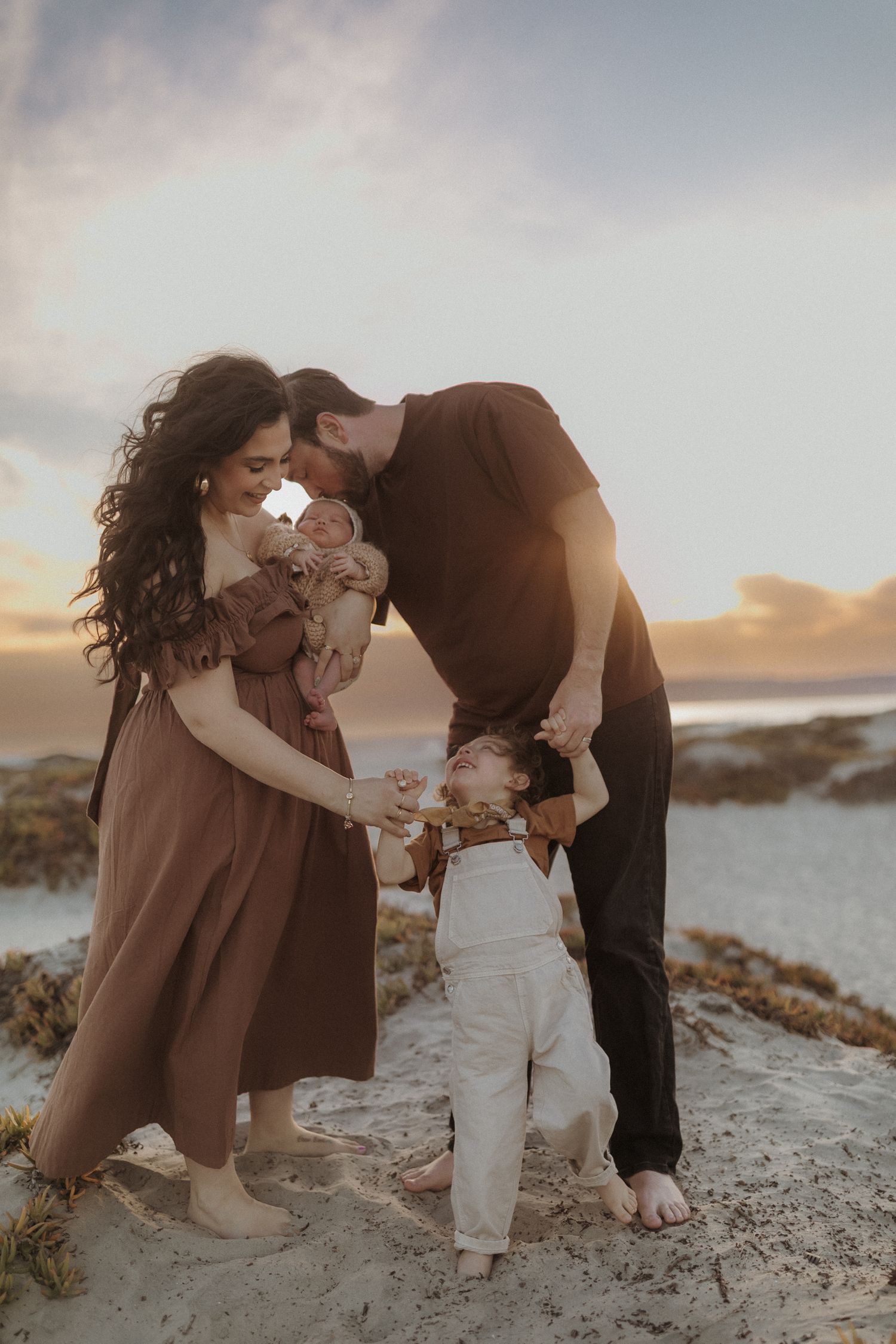 Coronado Newborn Photography. A family of three shares tender moments on a sandy beach during golden hour sunset.