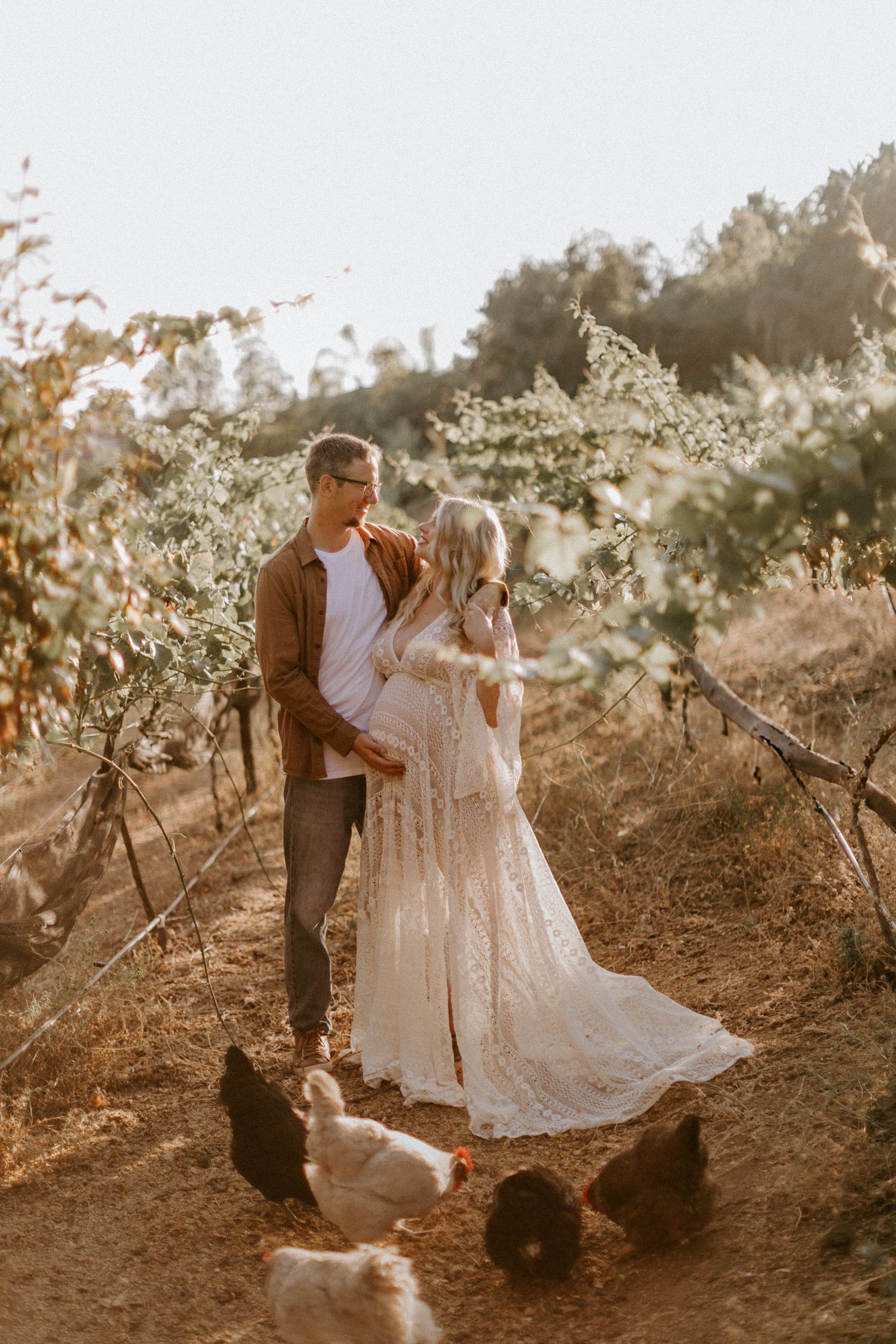 A couple stands in a sunlit San Diego orchard with chickens at their feet during a romantic outdoor maternity photoshoot at golden hour.