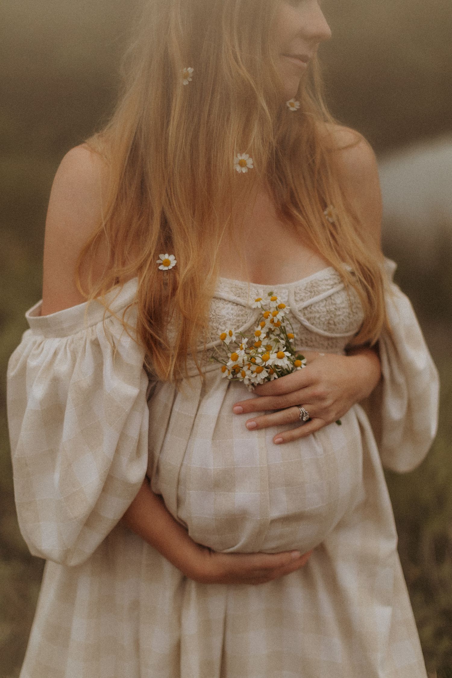 Woman in off-shoulder white dress holding wildflowers in golden light. Boho maternity photo shoot.