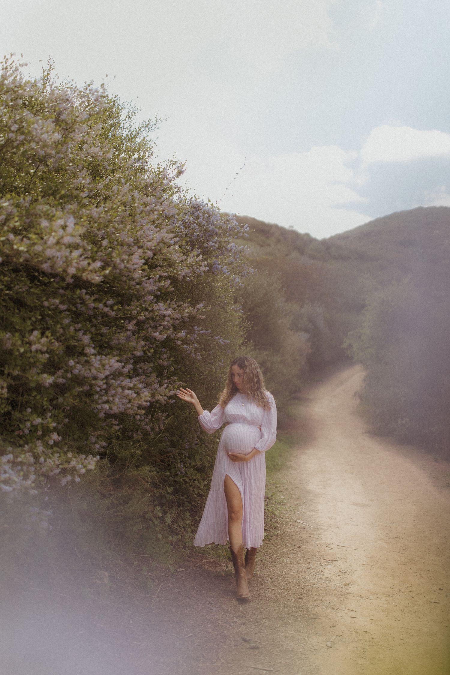 Dreamy figure in flowing dress walks through sunlit natural path. San Diego Maternity Photography.