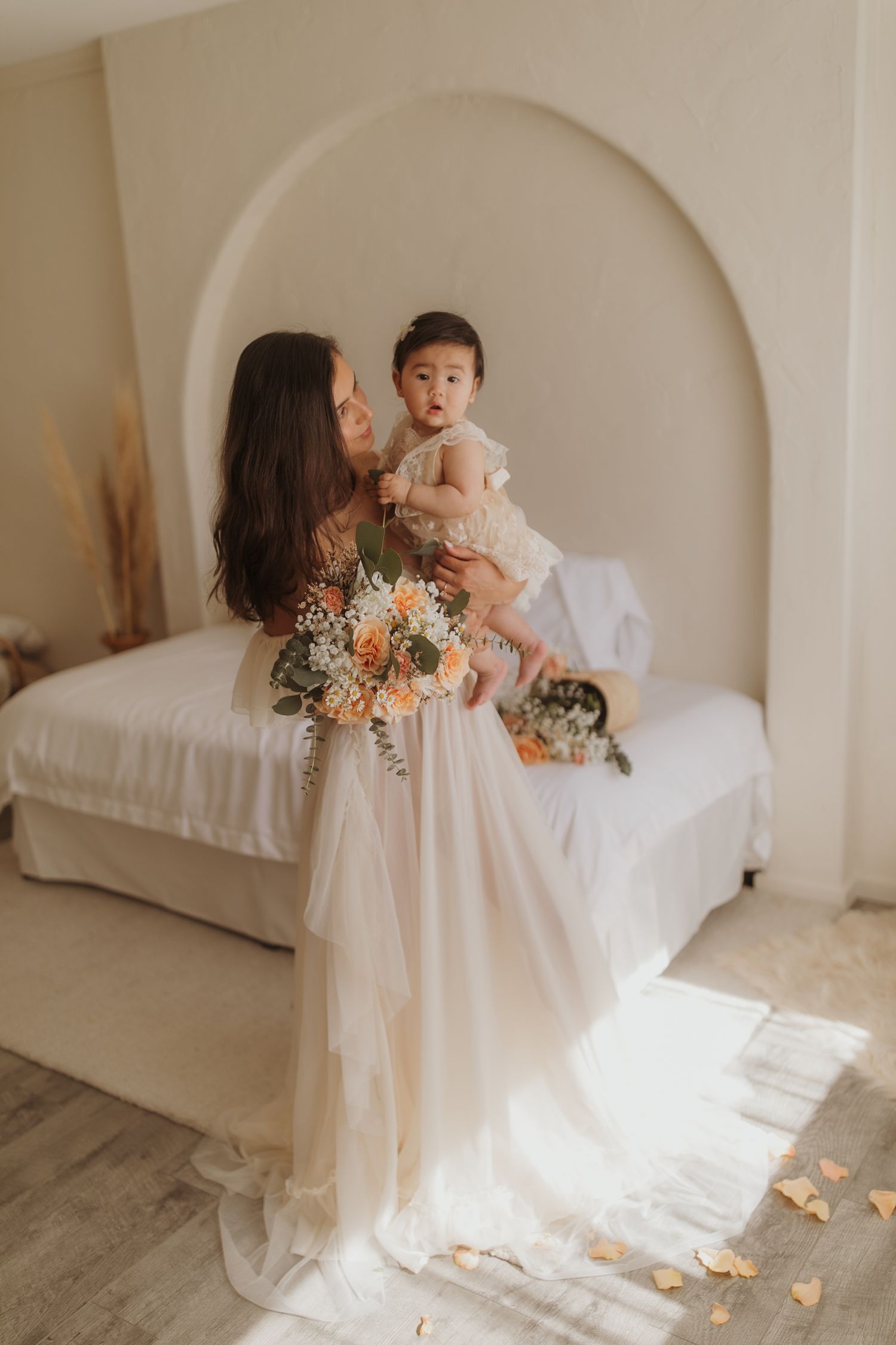 A tender moment between a mother and baby captured  in a minimal white bedroom with a flowing dress and delicate floral bouquet.