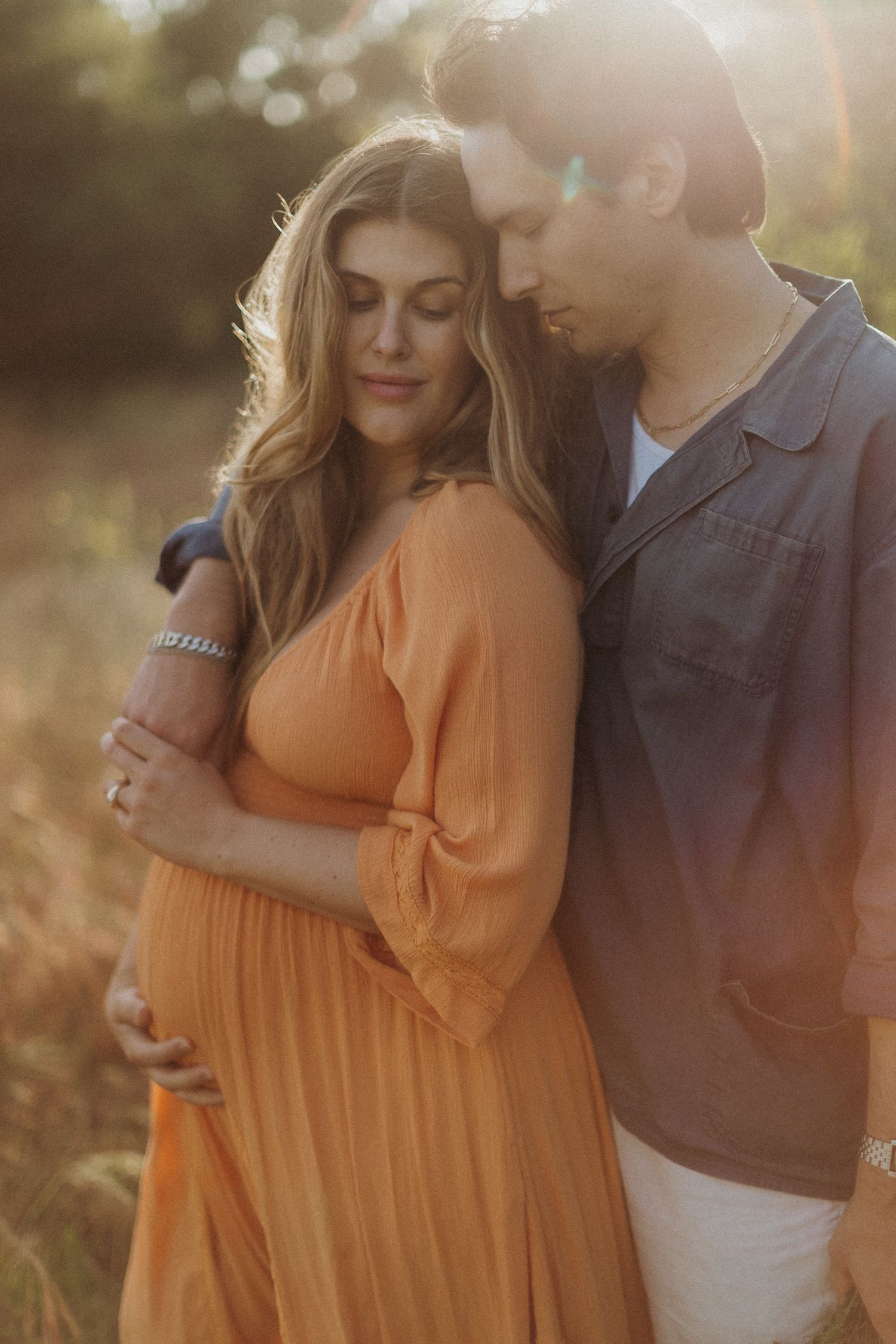 Couple embraces in golden sunlight while wearing orange dress and brown shirt in outdoor setting. San Diego Maternity Photographer.