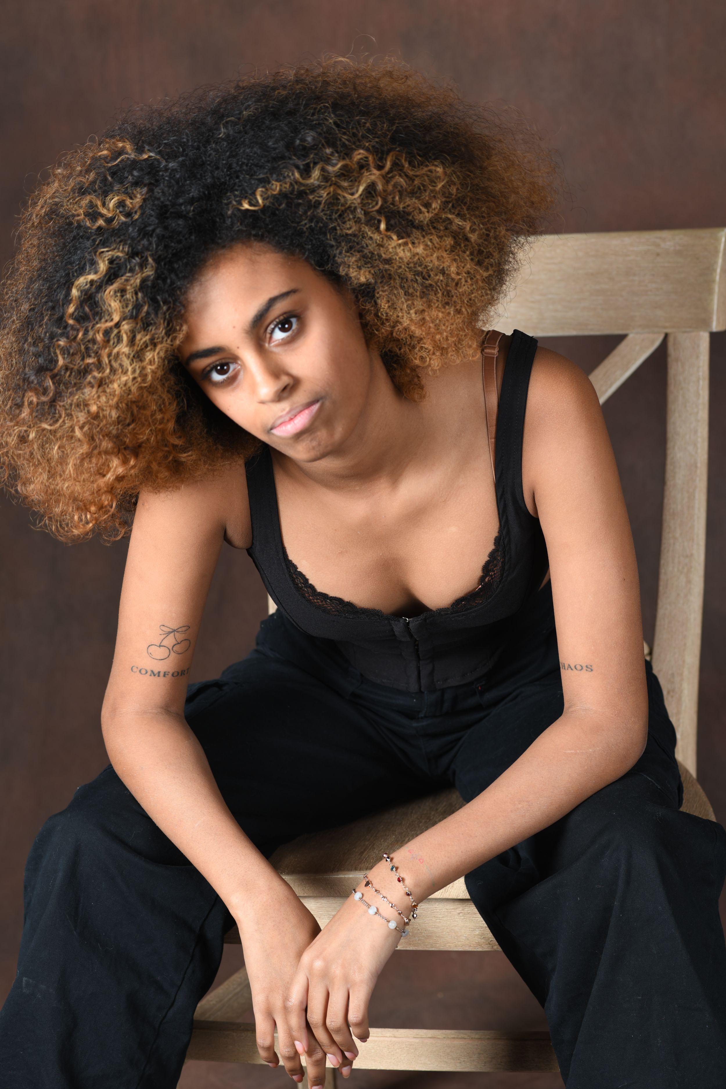 Portrait of person with curly highlighted hair wearing black top while sitting on wooden chair.