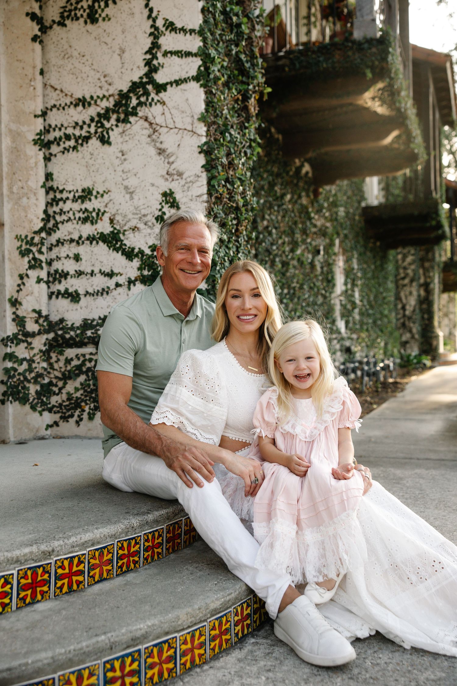 Family of three sitting on decorative tiled steps in front of ivy-covered wall, smiling warmly.