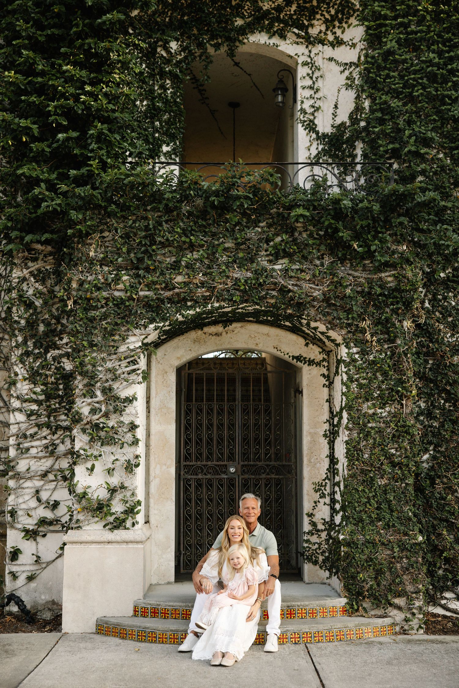Family of three posing on ivy-covered building steps with ornate iron gate backdrop.