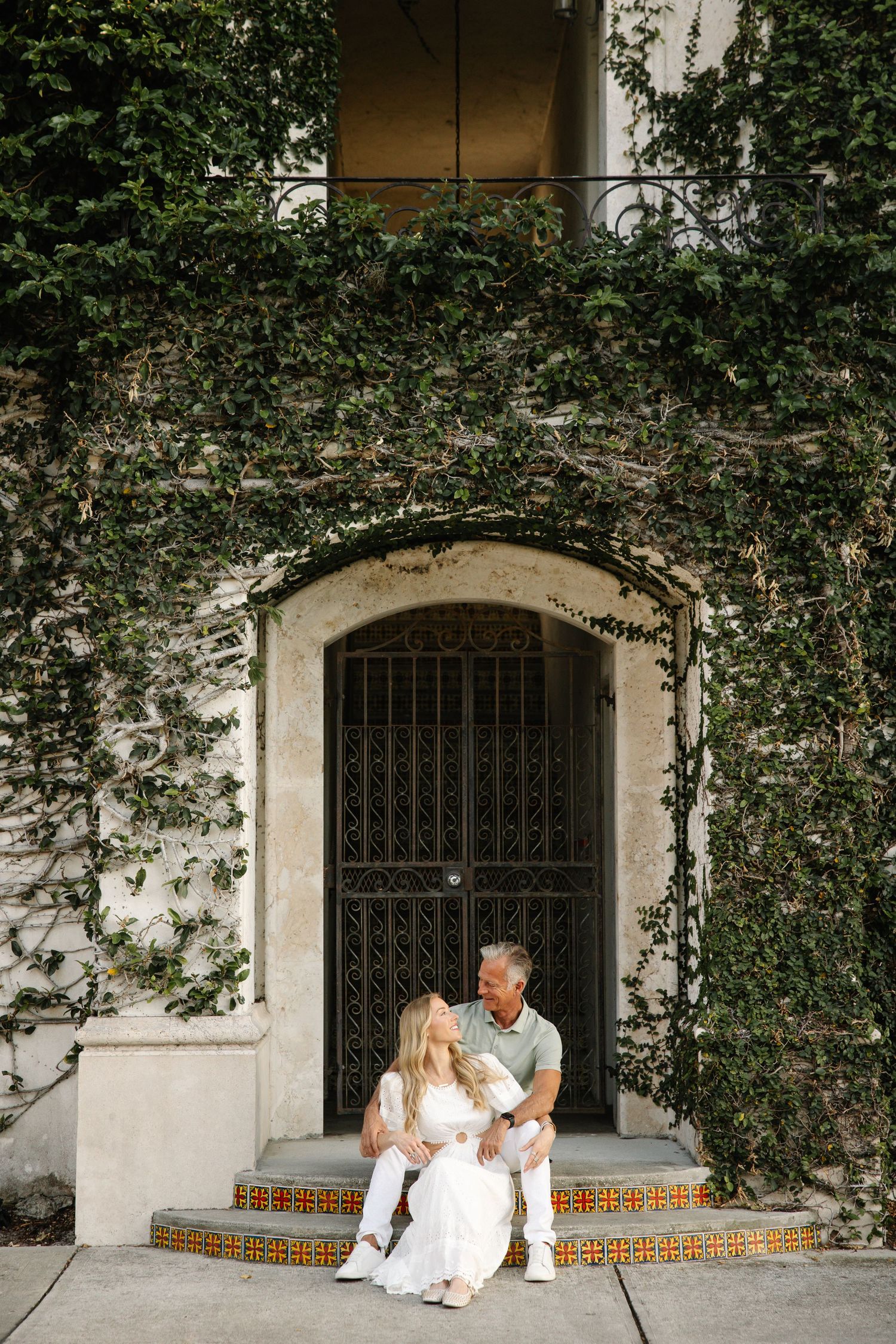 Couple sitting on tiled steps in front of ivy-covered building with arched iron door, dressed in white.