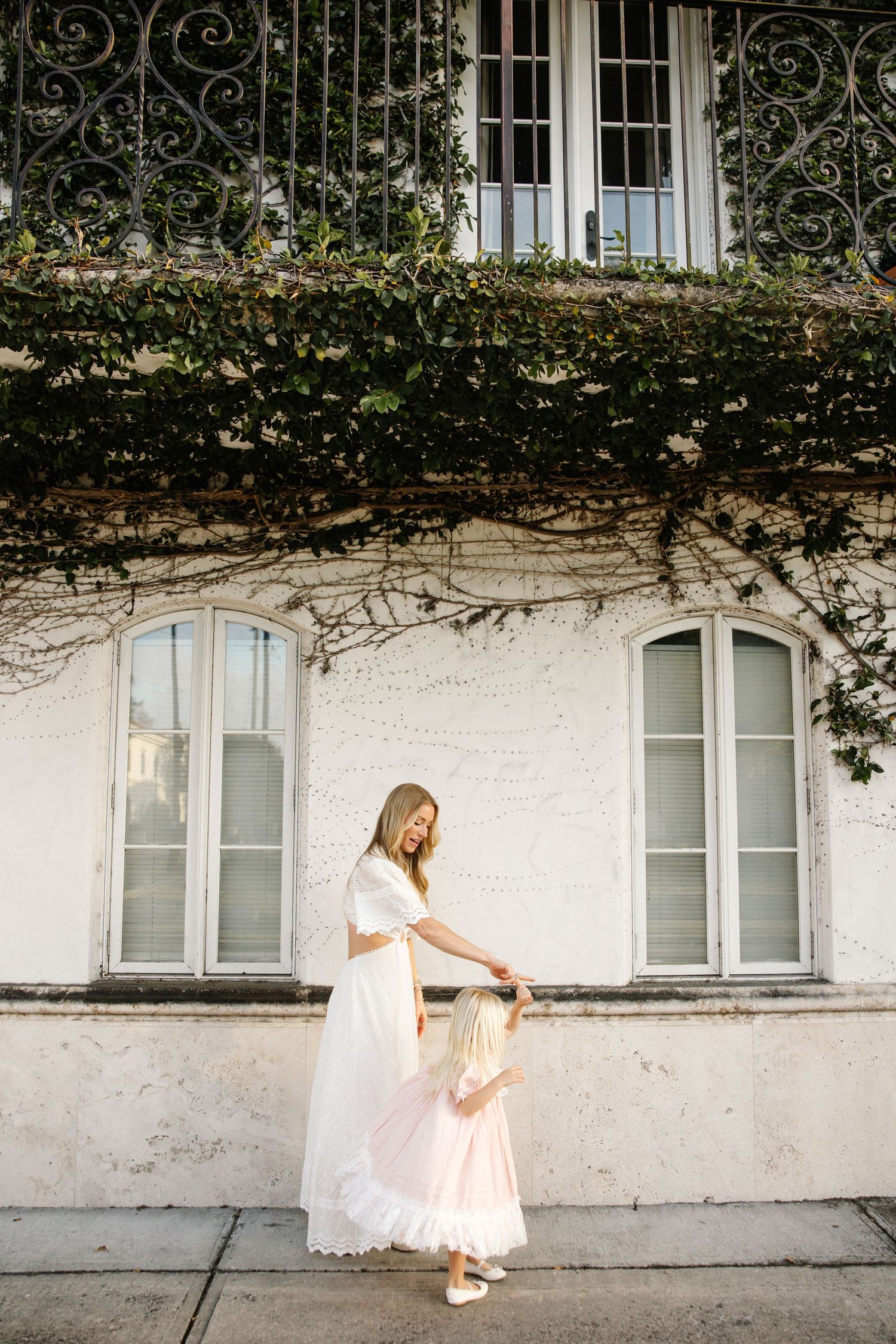 Woman in white dress holding child's hand outside ivy-covered building with arched windows.