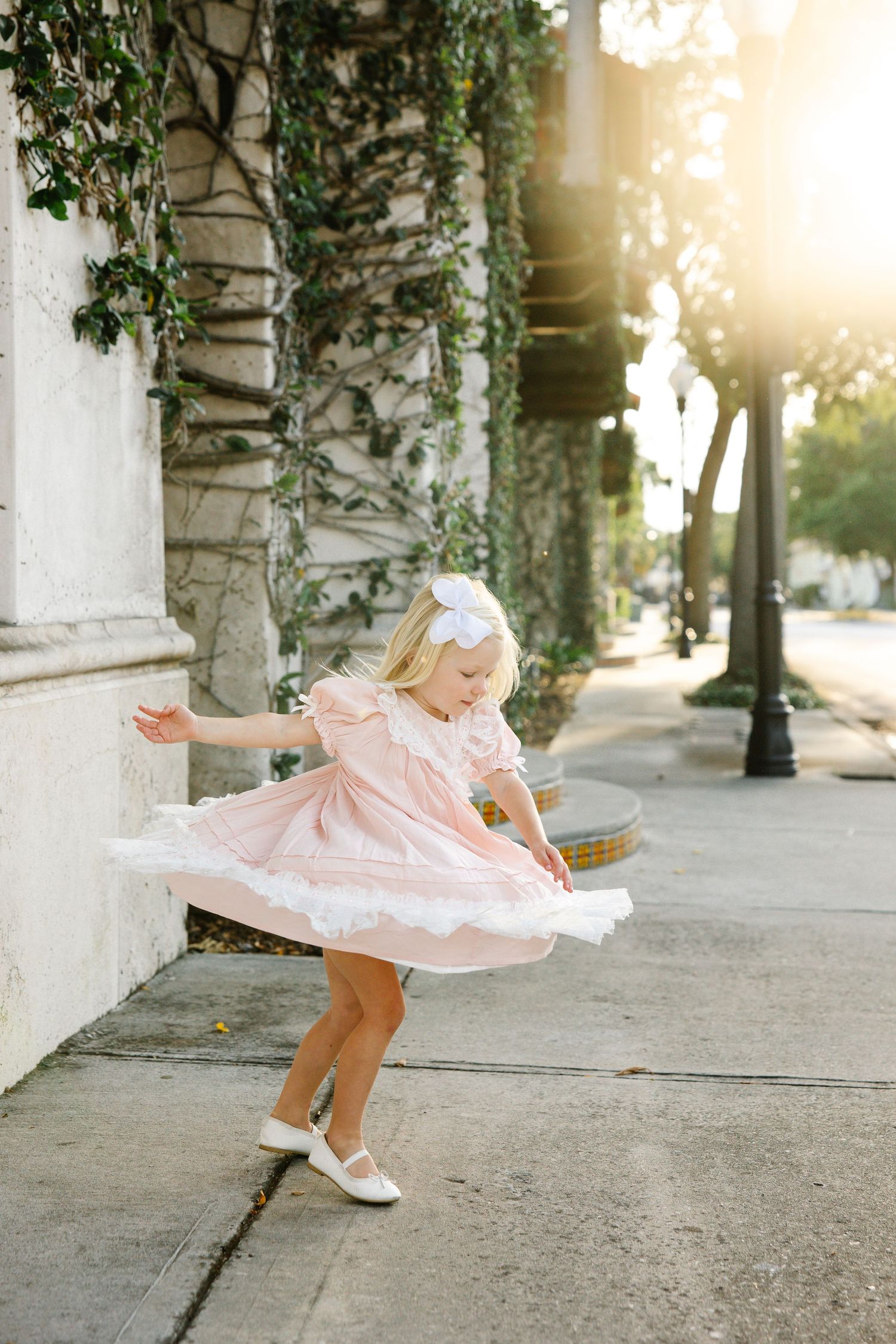 Young girl in pink dress twirling on a sunlit sidewalk, wearing white bow and shoes.
