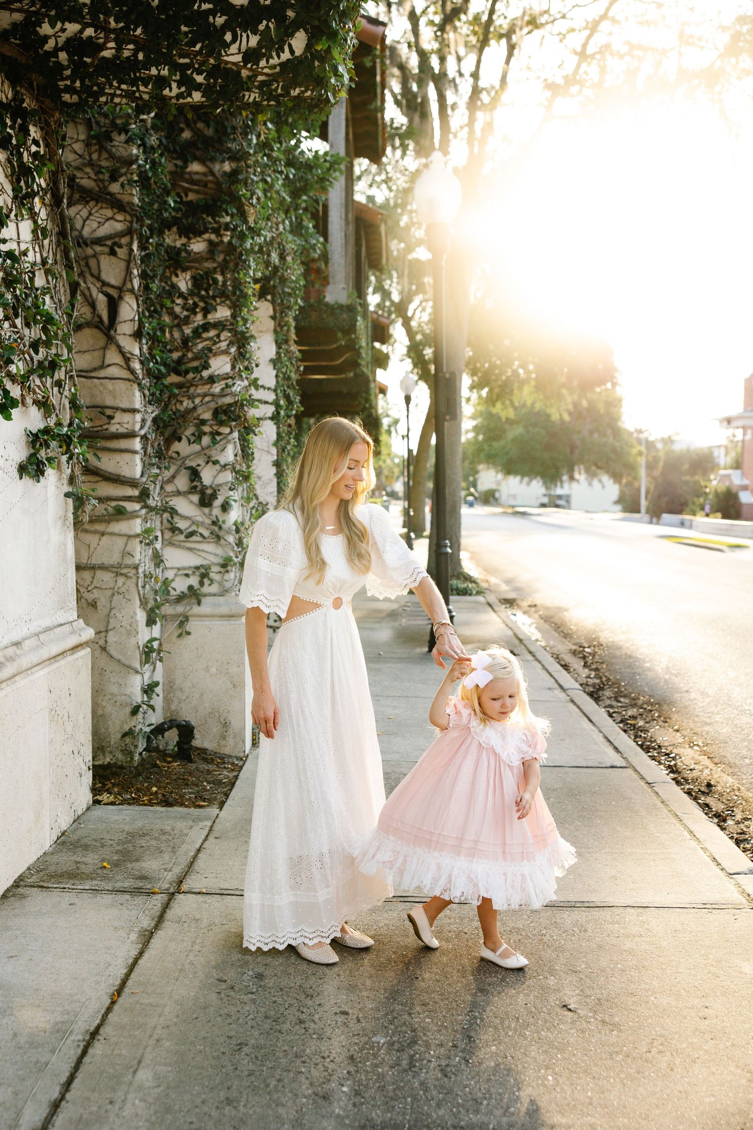 Mother in white dress walks with toddler girl in pink dress on sunny sidewalk.
