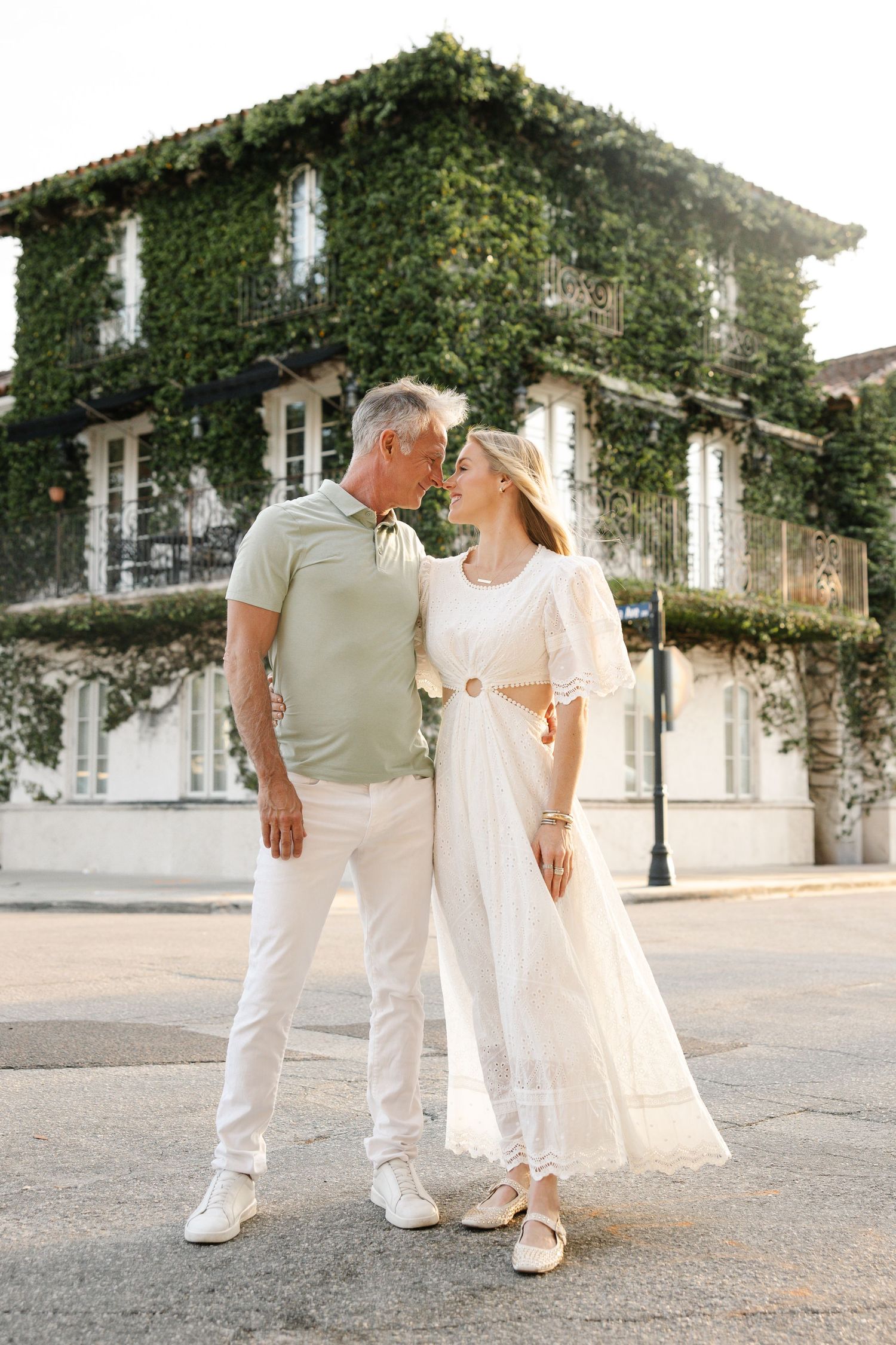 Couple in white and sage outfits sharing a tender moment outside ivy-covered building.