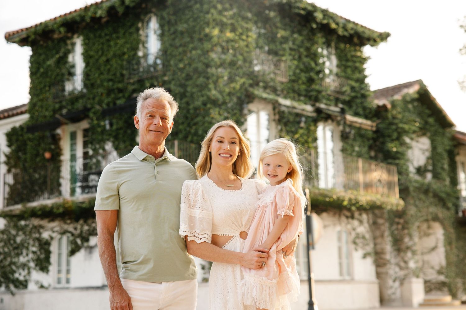 Family of three smiling outdoors in front of an ivy-covered house, parents holding young blonde girl.