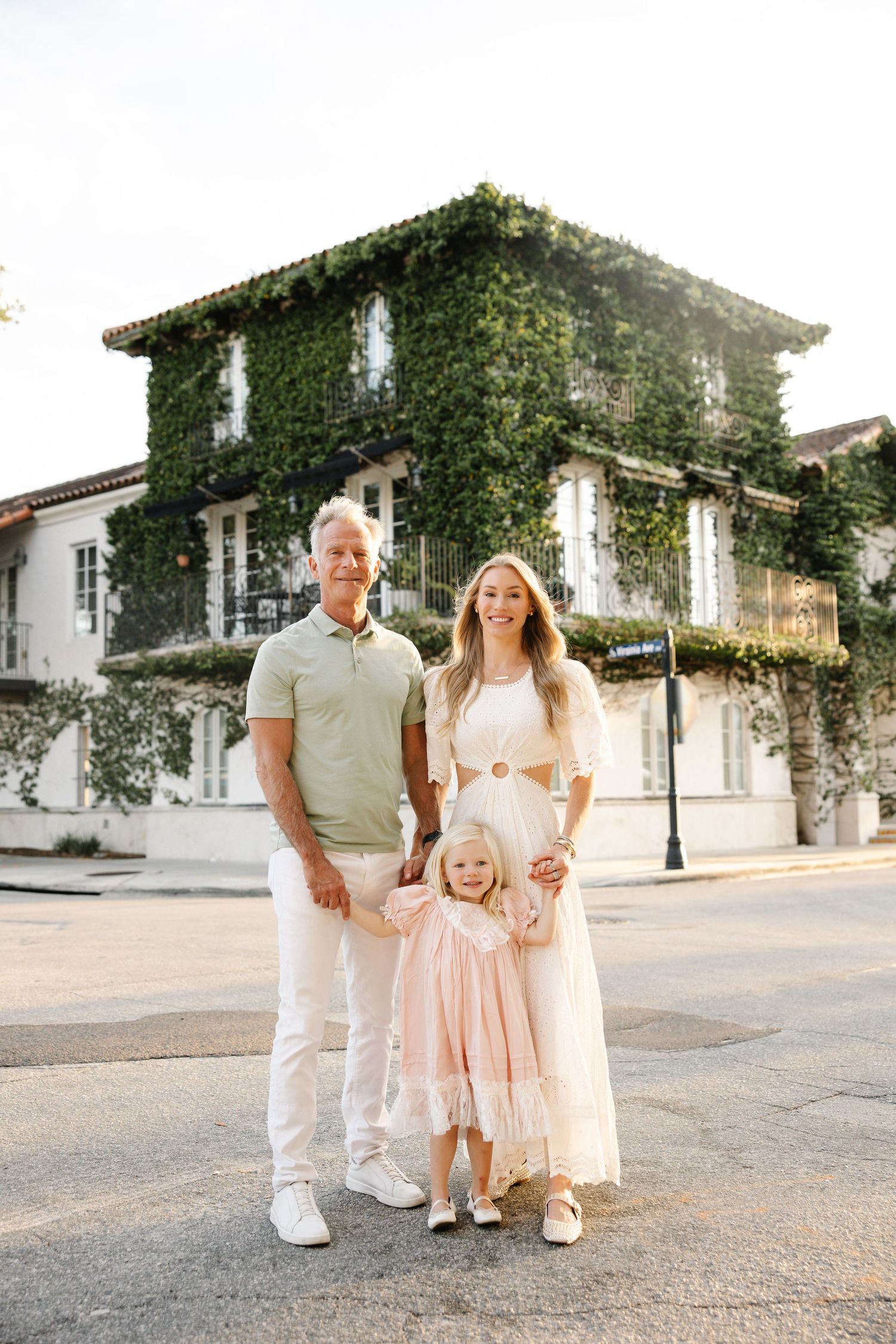 Family of three posing in front of ivy-covered white building, parents in neutral tones, toddler in pink dress.