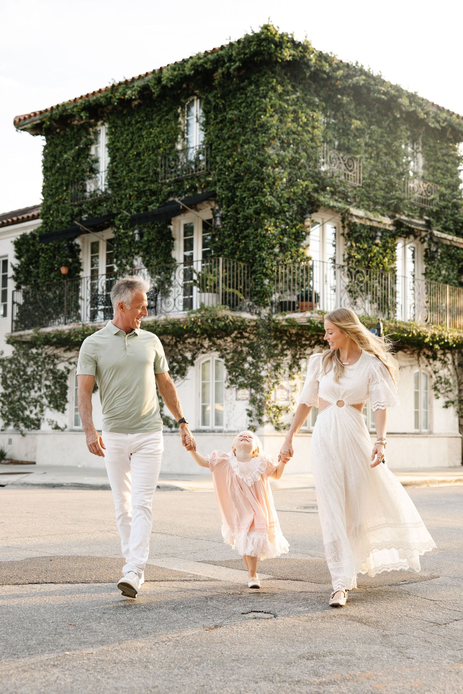 A happy family with a toddler girl walks hand-in-hand outside an ivy-covered building in warm sunlight.