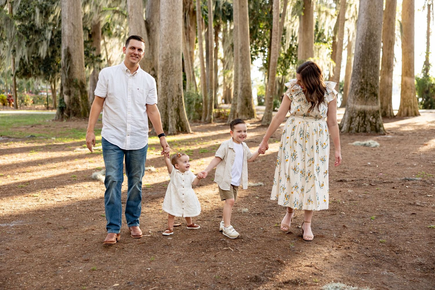 A playful family photo session in a sunlit wooded park with towering trees and natural lighting.