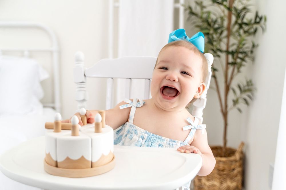 Adorable baby girl in a blue bow laughing joyfully in front of a wooden birthday cake.