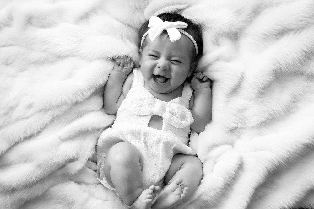 Black and white photo of joyful baby in white outfit with headband laying on blanket.