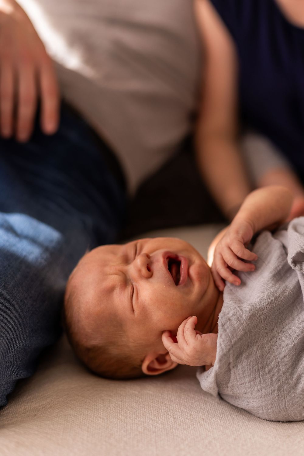 A crying baby in white clothing lies on a light colored couch.