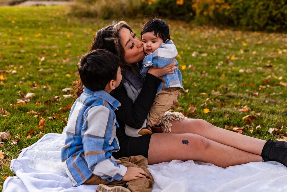 A family shares a joyful moment together while sitting on a blanket in a park with fallen autumn leaves scattered around.