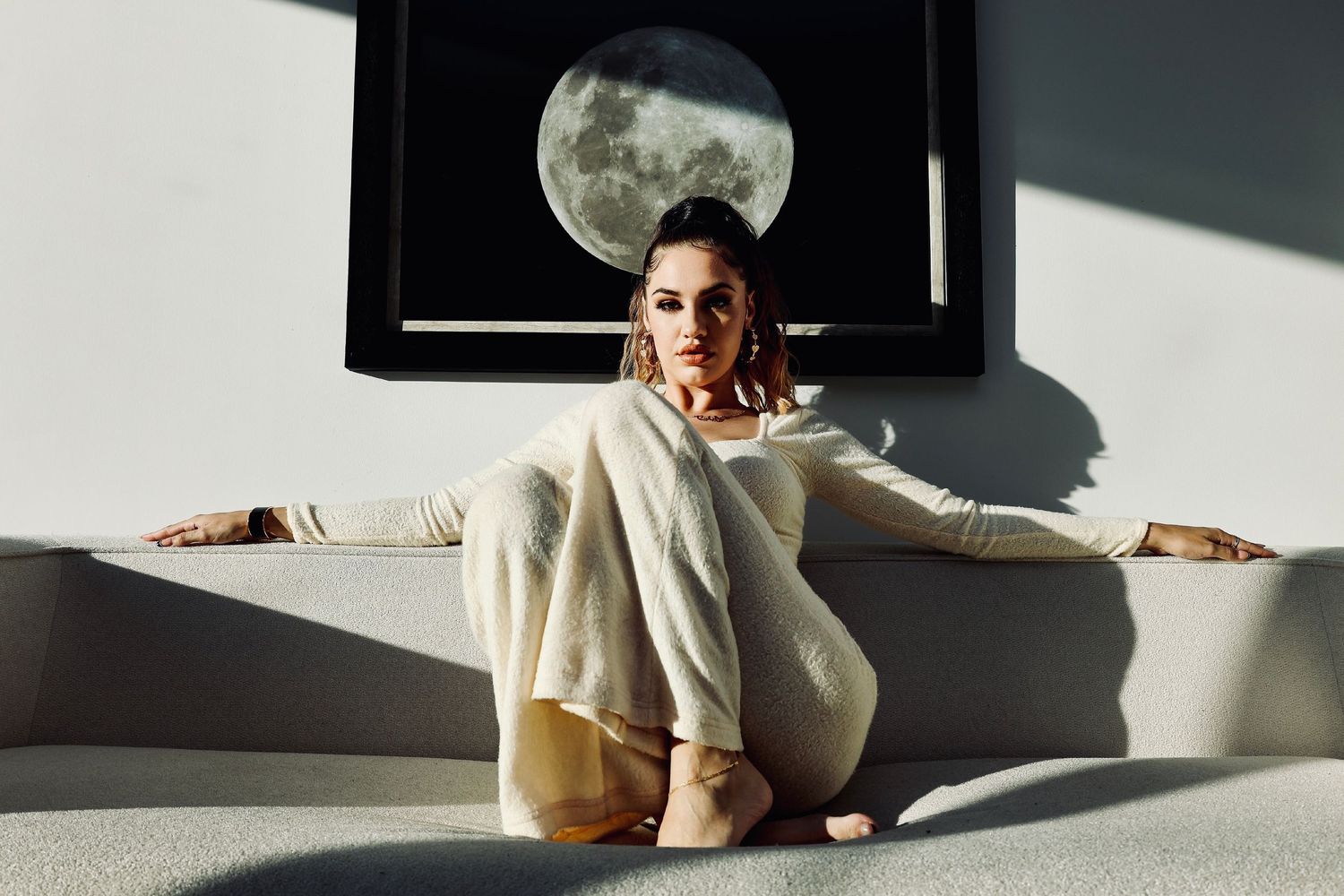 Model in cream sweater sitting beneath framed moon artwork in dramatic lighting and shadows.