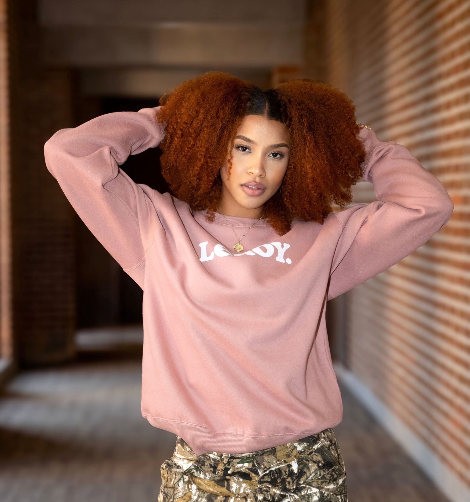 Model in pink sweatshirt poses casually in hallway setting.
