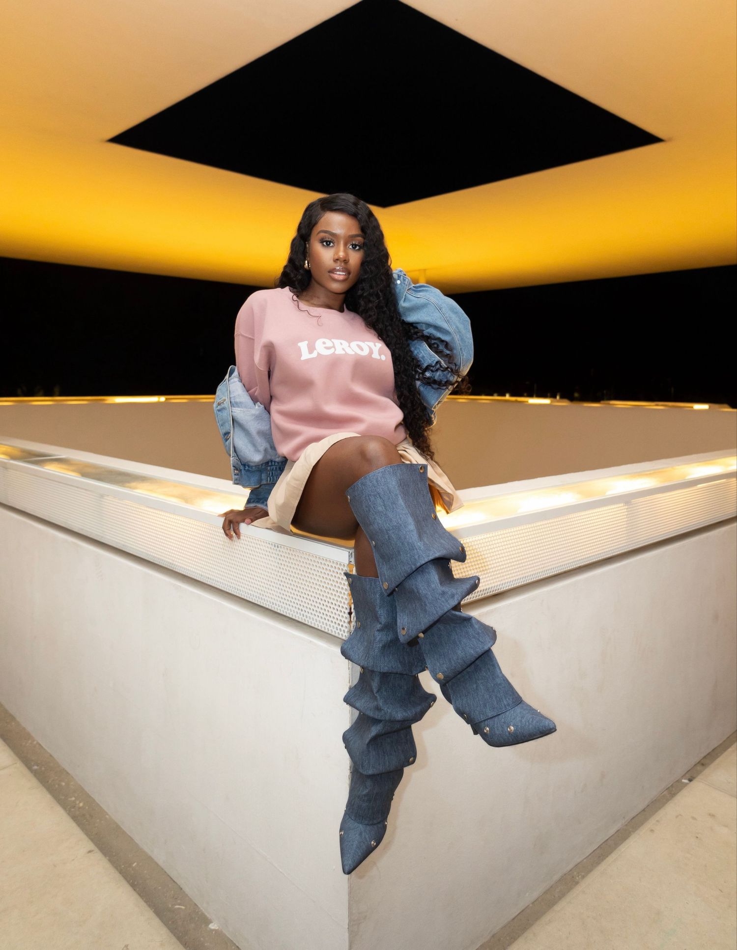 Fashion photo with model in pink sweatshirt and stacked denim boots against illuminated backdrop.