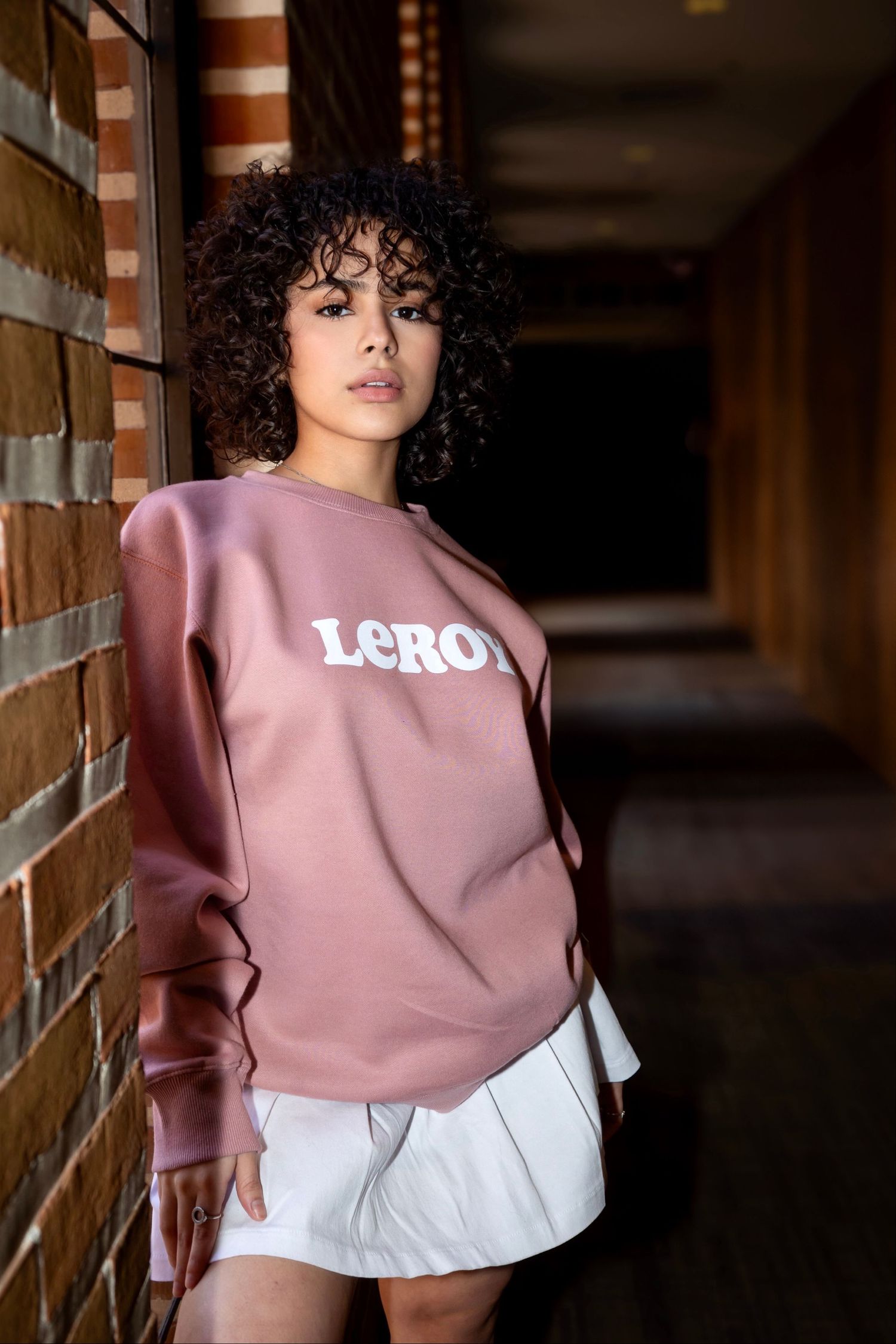 Pink LCRD sweatshirt and white skirt photographed in a dimly lit hallway with brick walls.