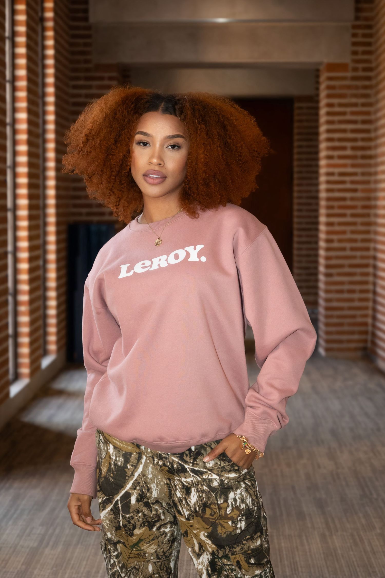 Fashion model wearing pink Lerox sweatshirt and camo pants in indoor setting.