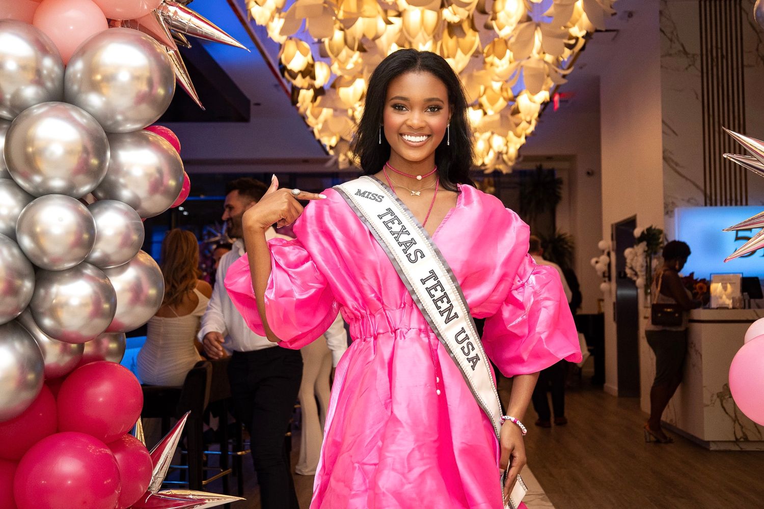 A pageant winner in a bright pink puffy dress poses with a crown and sash amid silver and pink balloon decorations.