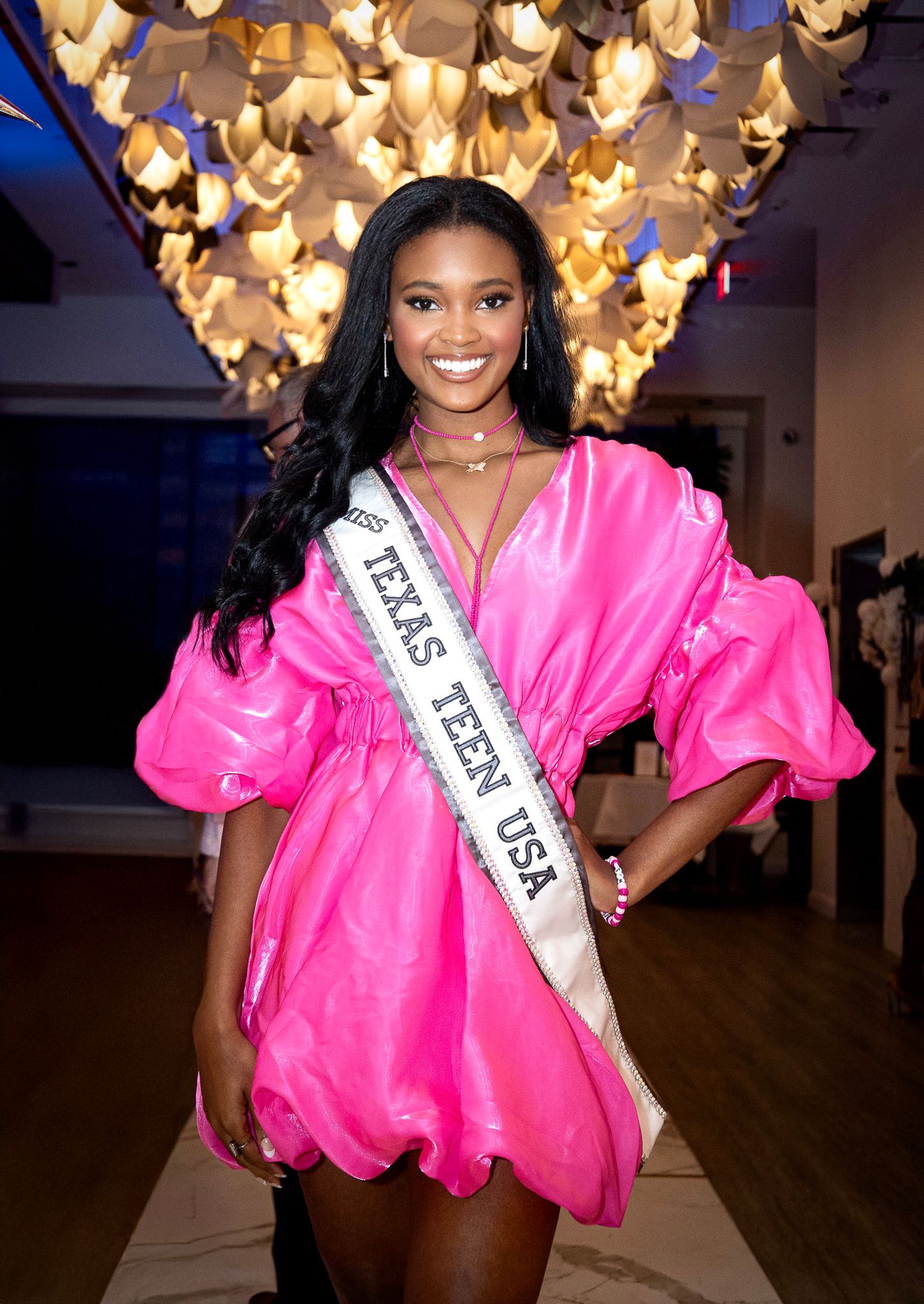 A pageant winner wearing a vibrant hot pink dress and Miss Teen USA sash poses under warm glowing lights.