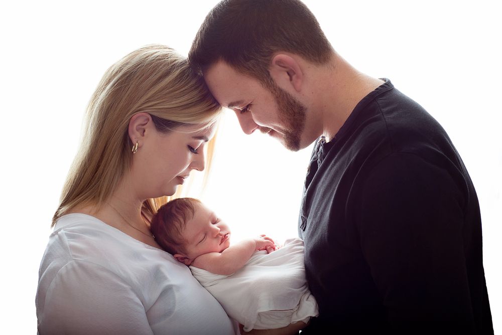 backlit side profile close up of mom and dad with heads together  with newborn baby held between. in studio newborn session Baltimore, ON*