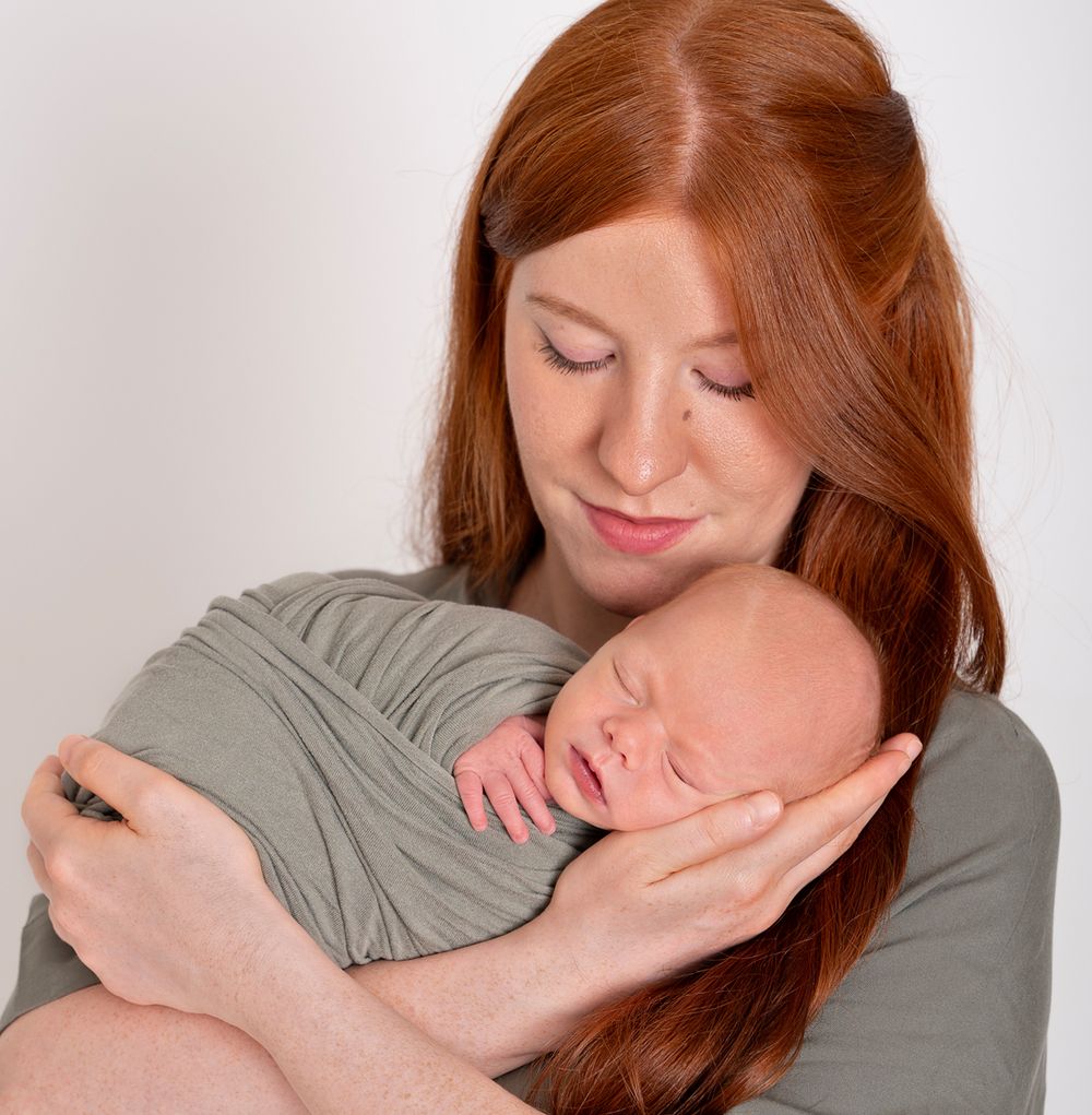 mother looking down gently embracing newborn baby boy in her arms wearing sage green dress and sage green wrap Baltimore Ontario*
