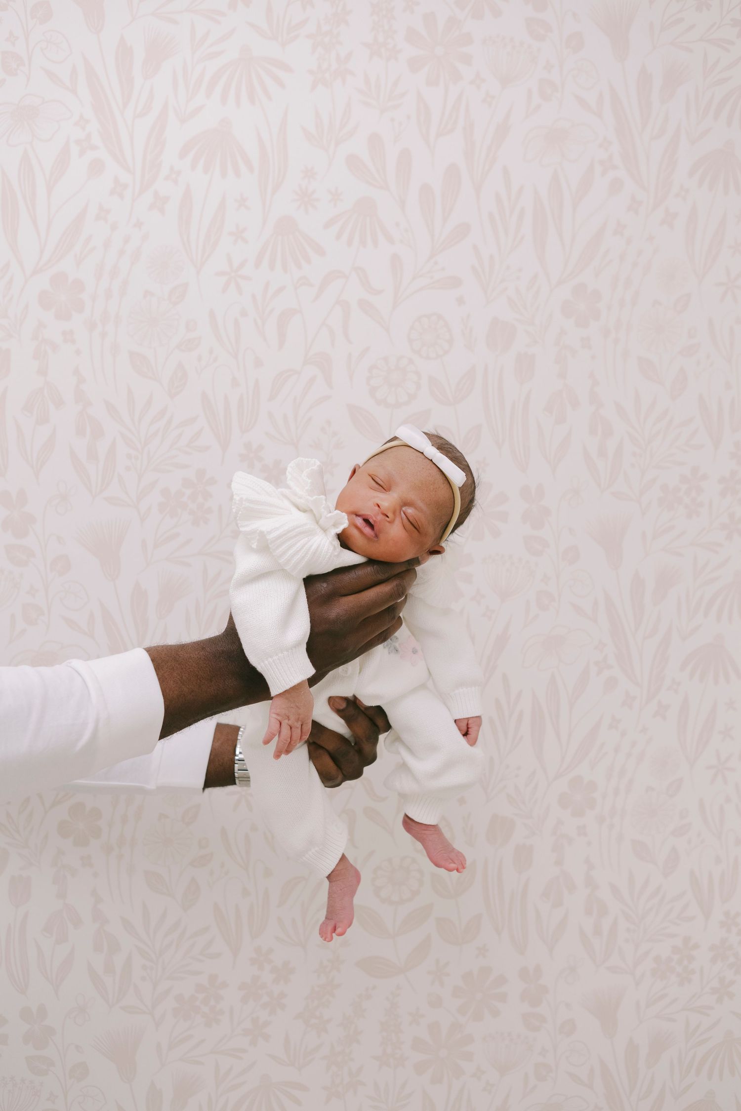 Hands of dad holding the baby against floral wallpaper, captured by Paula Ortiz during a family session