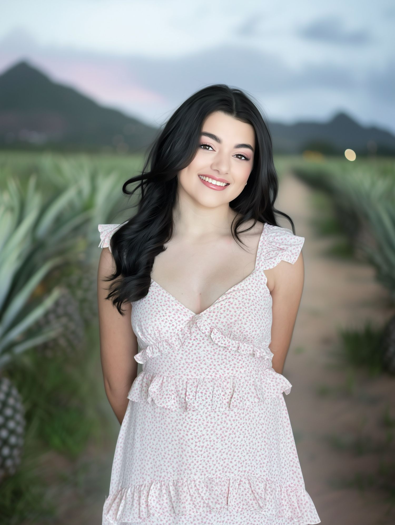 A person in a white ruffled sundress poses on a path through a pineapple field with mountains in the background.