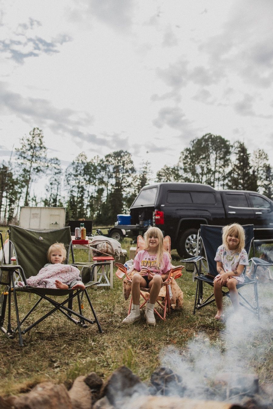 Three children sit in camping chairs around a campfire at a forest campsite with a black truck parked nearby.