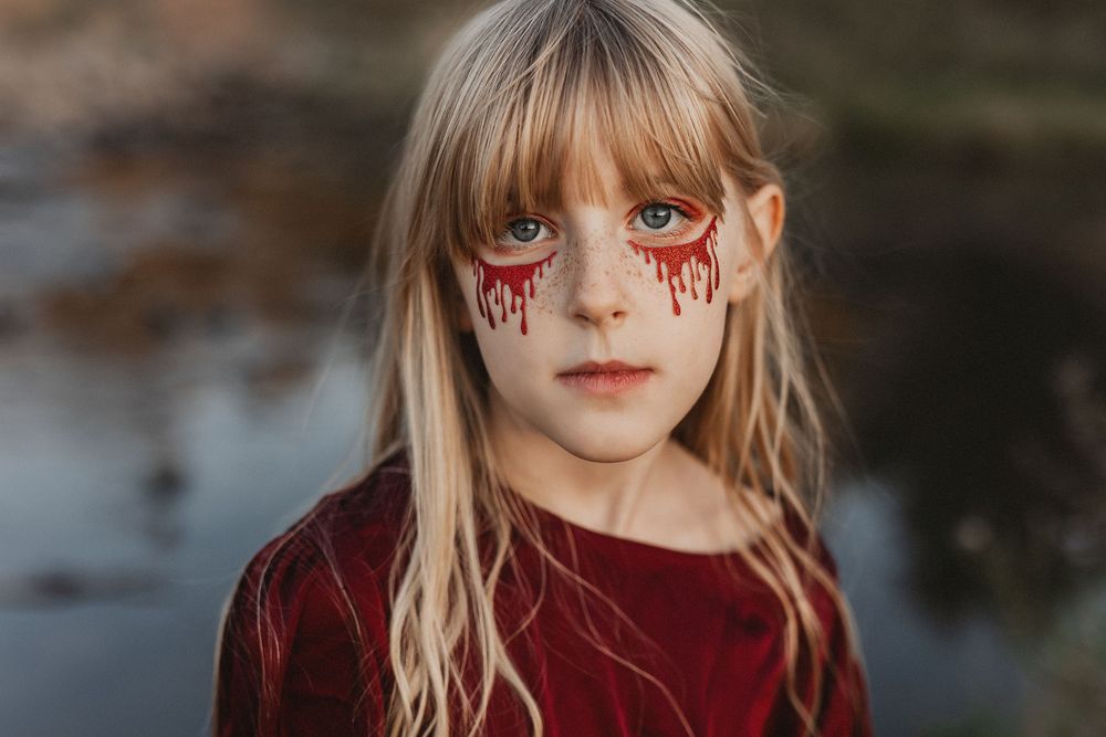 Young girl with blonde hair and Halloween makeup featuring red dripping tears on her cheeks, wearing a red top.